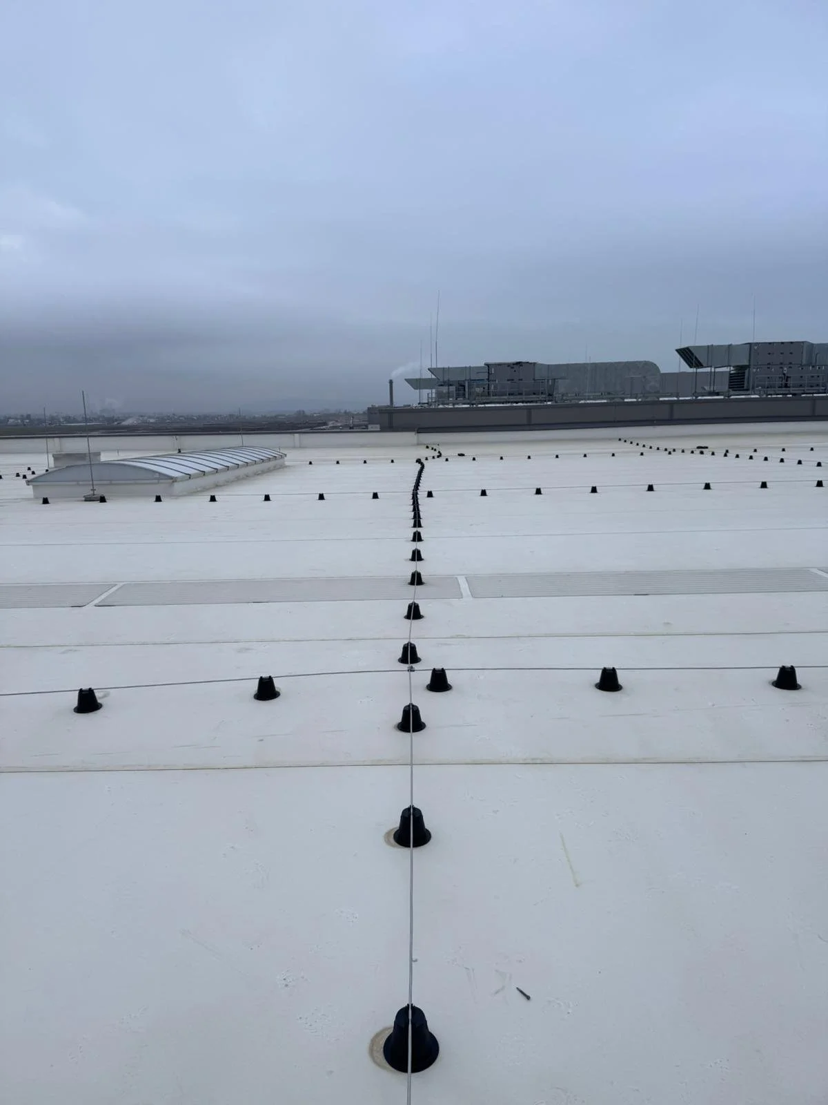 View of a white building rooftop with black safety cable anchors and vents, under a cloudy sky.