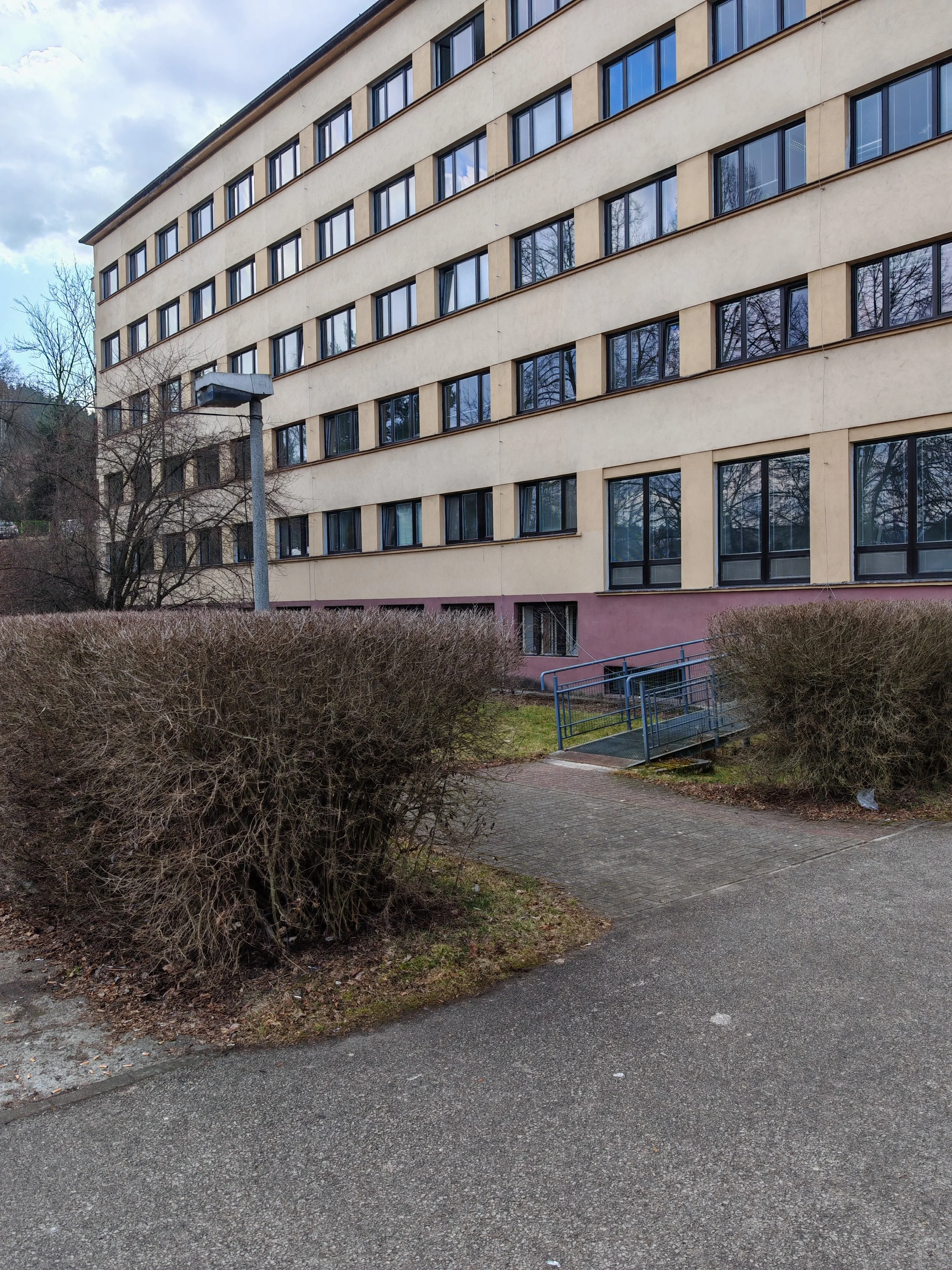 A multi-story beige apartment building with large windows, a sloped roof, and a small entrance stairway, set against a cloudy sky with bare trees and bushes in the foreground.