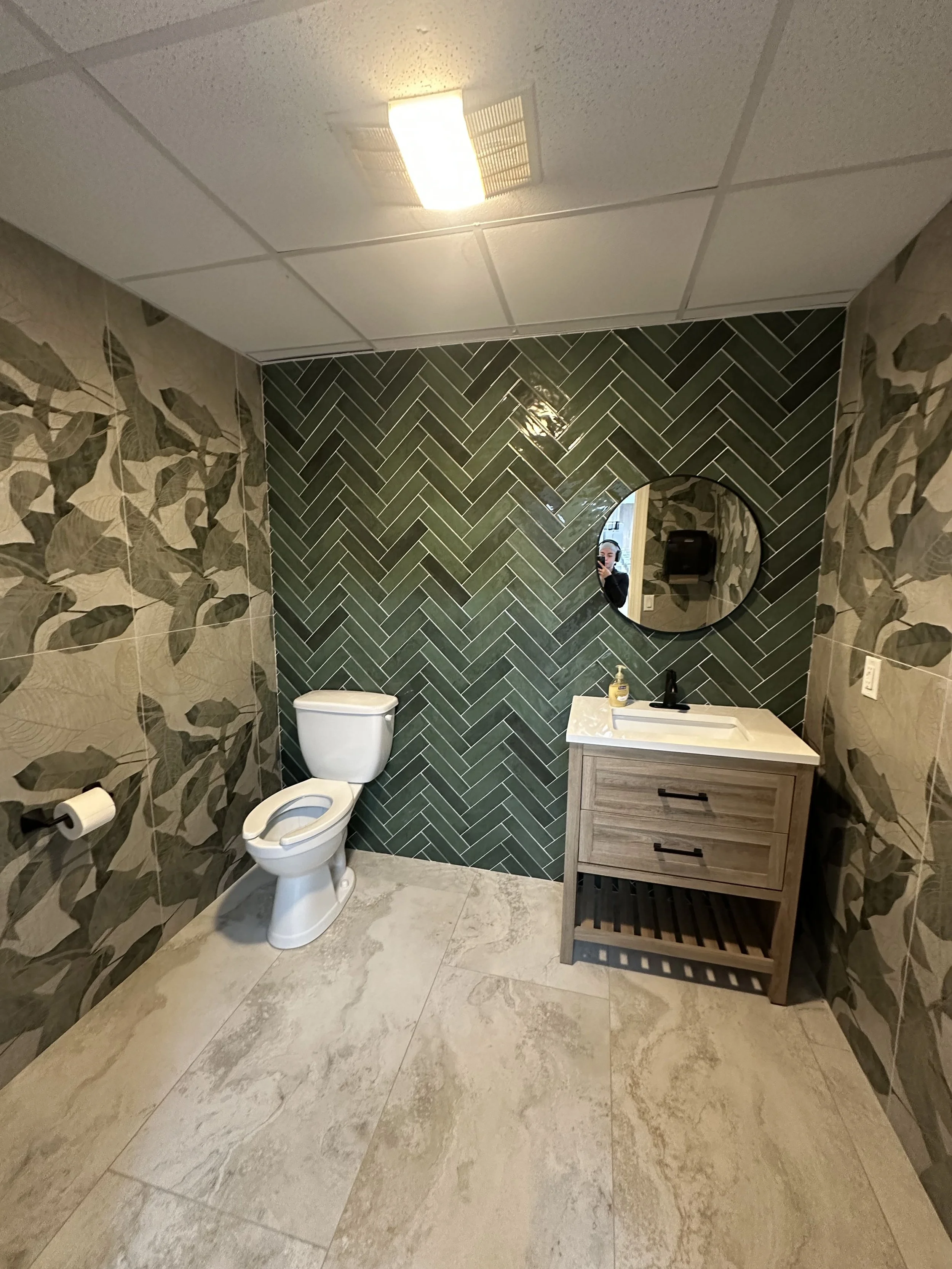 A bathroom with beige marble-like tile flooring, a white toilet, a small wooden vanity with a white sink and an oval mirror above it, a bottle of soap, a black faucet, a wall-mounted paper towel dispenser, and wallpaper with large leaves on the side walls. The back wall has dark green herringbone tiles.
