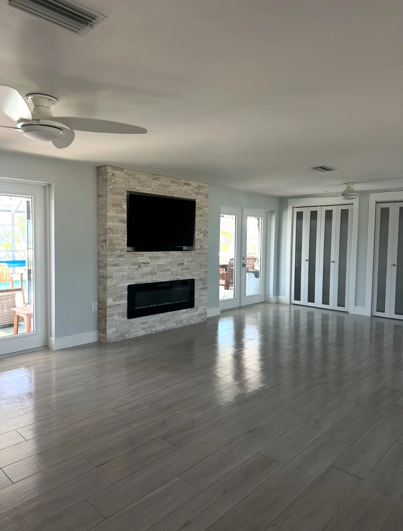 Modern living room with light wood flooring, stone accent wall, mounted TV and fireplace, ceiling fan, large windows, and glass doors leading outside.