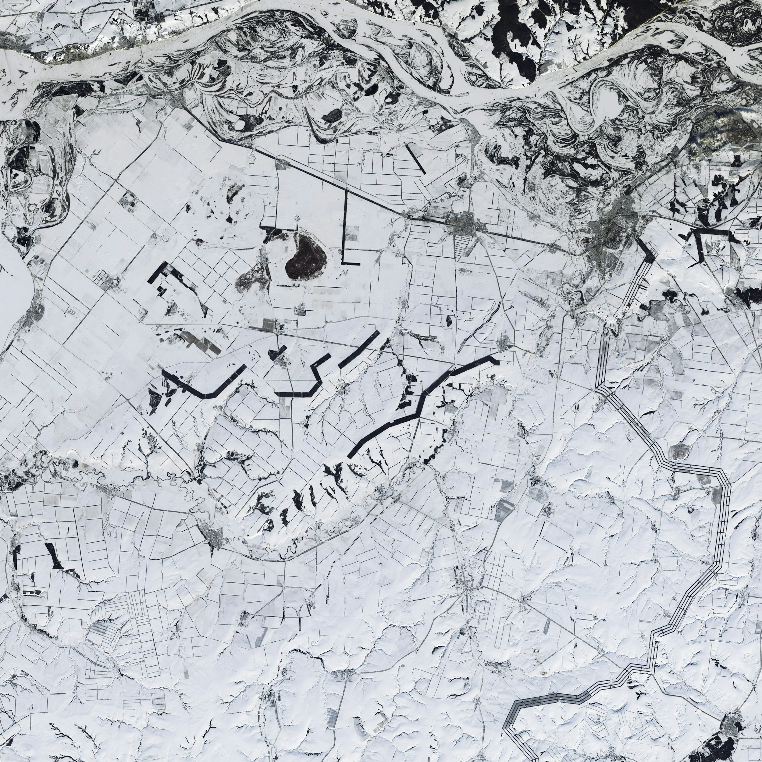 A top-down aerial view of a snow-covered landscape with roads and farmland, featuring snow-capped hills and a network of black lines representing roads or pathways.