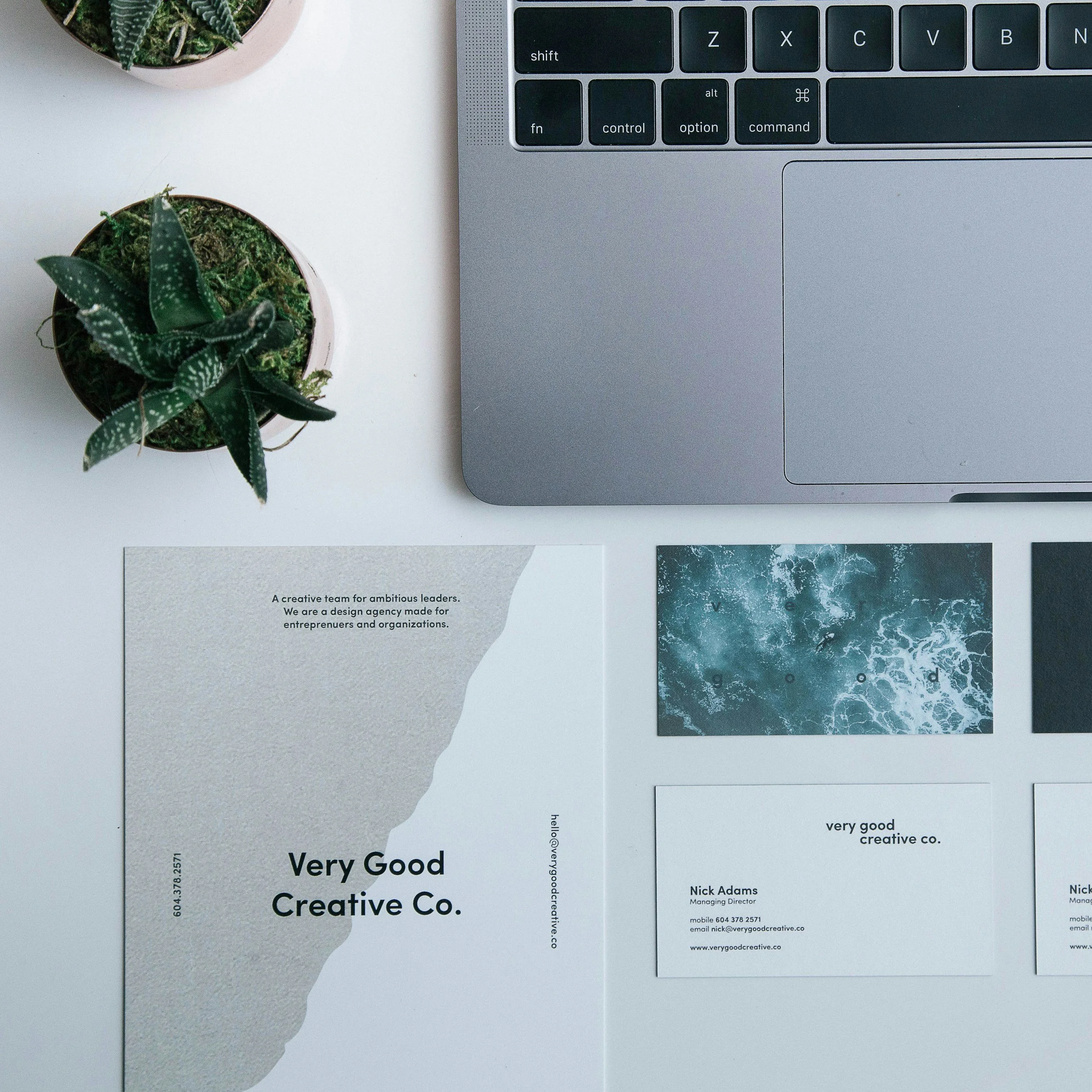 Flat lay of a workspace with a laptop, two small potted plants, a business card, and printed materials from a creative company on a white surface.