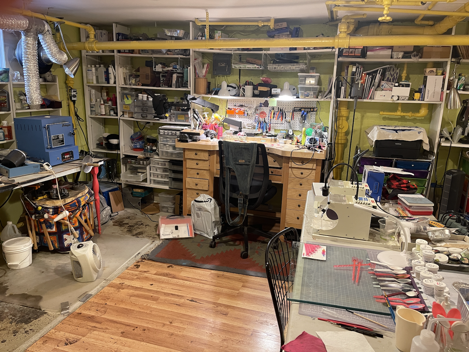 A cluttered workshop with shelves filled with tools, supplies, and various equipment. There is a wooden workbench with a chair in front, and a busy table with craft or art supplies. The floor is a combination of hardwood and concrete, with some areas showing signs of use and dirt. Yellow pipes run along the ceiling and wall, and various lamps and devices are scattered throughout the space.