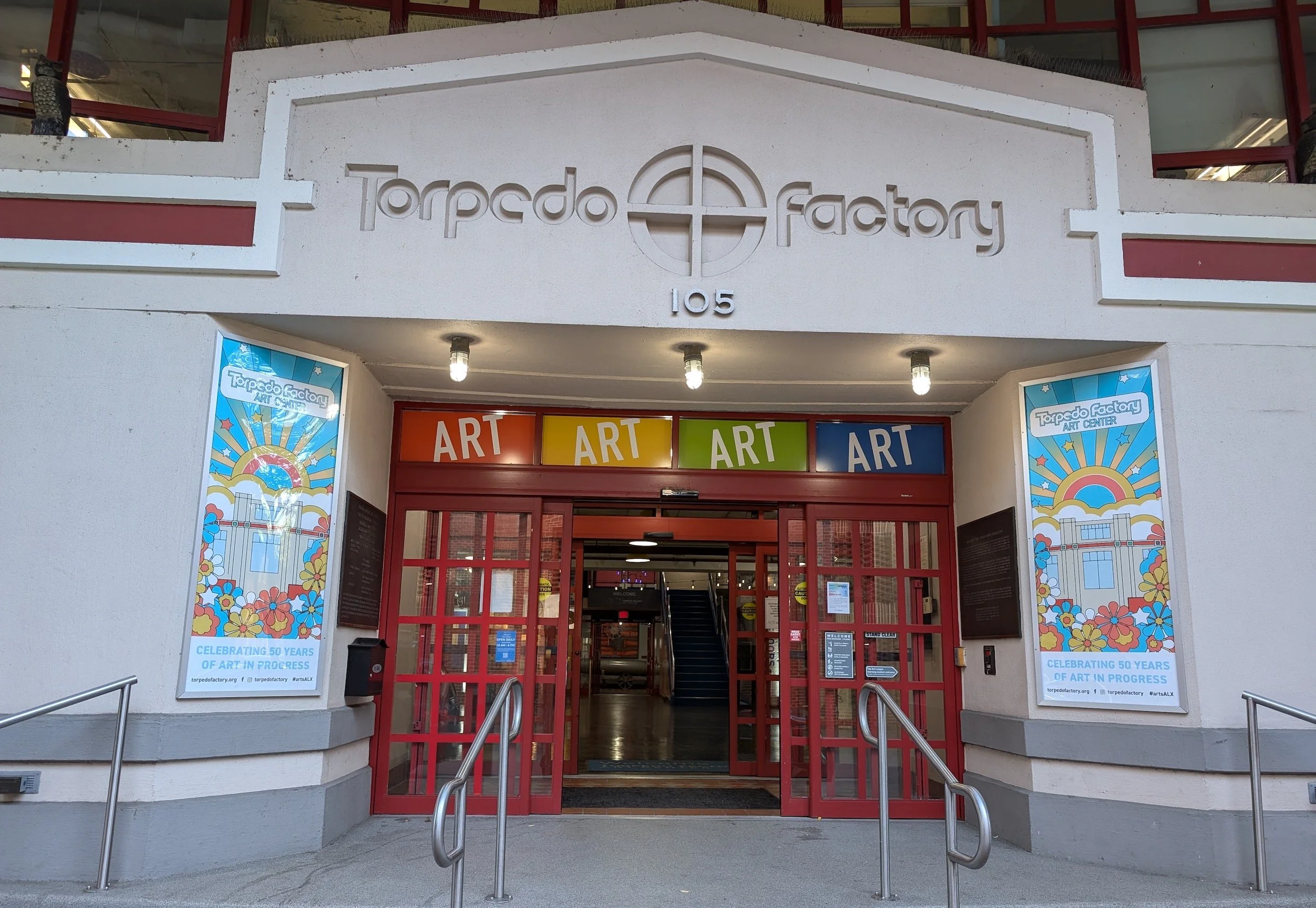 Entrance to the Torpedo Factory Art Center, with colorful ART signs above the door, and two poster advertisements on either side celebrating 50 years of art in progress.