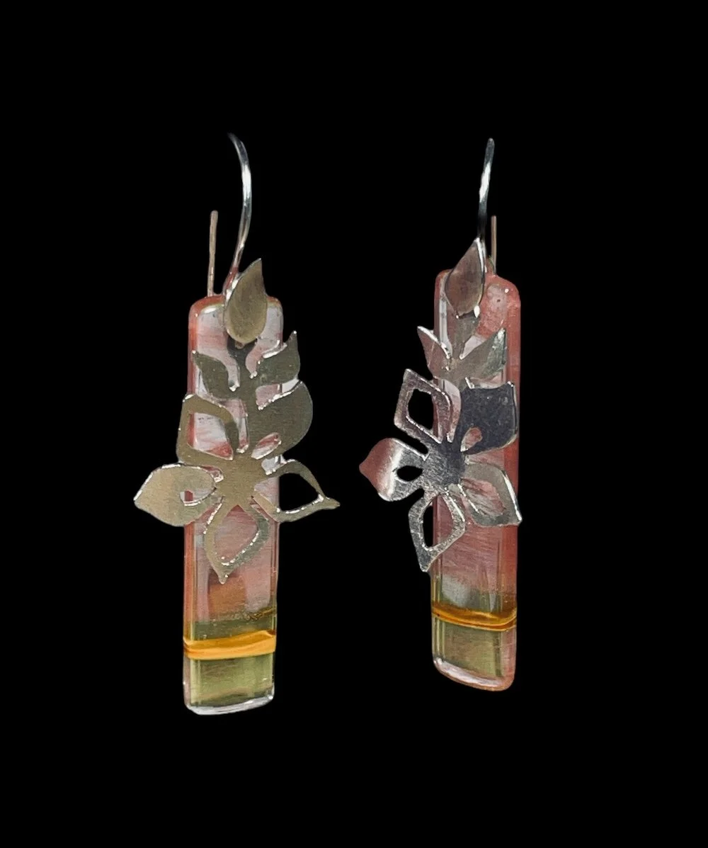 Silver earrings with leaf and floral cut-out designs, hanging from a pink and green rectangular glass base on a black background.