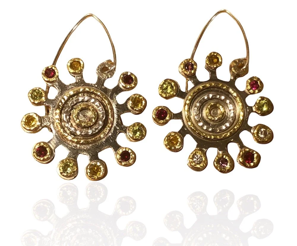 Gold and red gemstone earrings with circular design, featuring small stones around a central spiral pattern, reflected on a white surface.