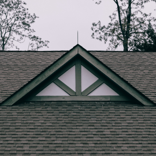 Close-up of a house roof gable with a decorative truss, surrounded by trees under an overcast sky.