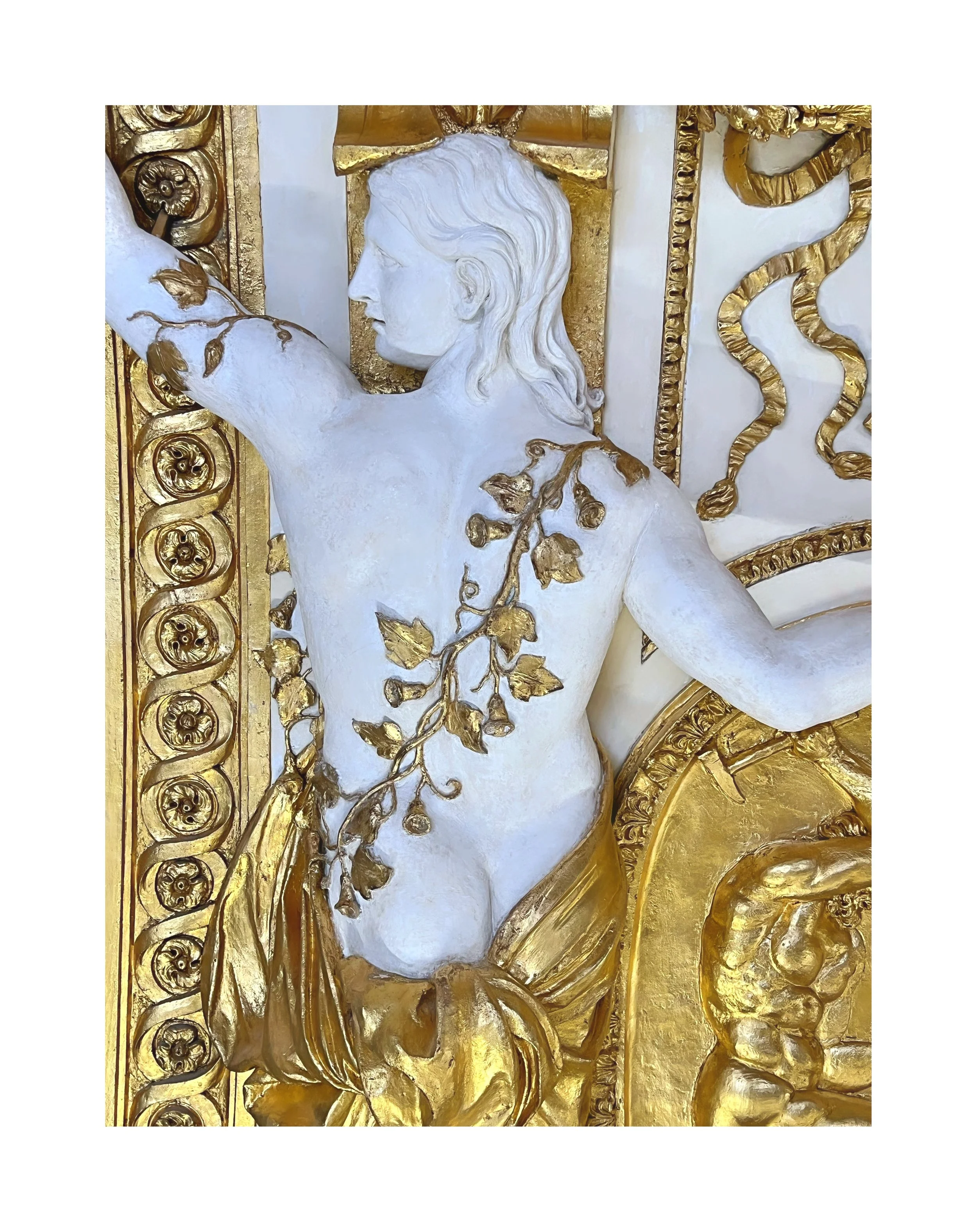 Sculpture en or et blanc représentant un homme nu avec une couronne de fleurs, dans un style classique, ornée de détails floraux et décoratifs.