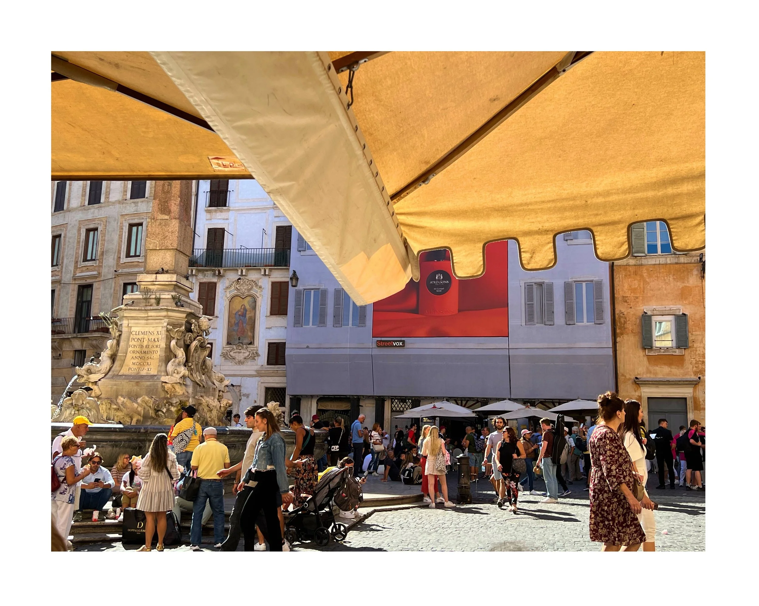 Une place publique animée avec de nombreuses personnes, des bancs, un monument avec une fontaine et des sculptures, des immeubles historiques en arrière-plan, et une grande publicité à l'écran. Ombres portées par un auvent jaune au premier plan.