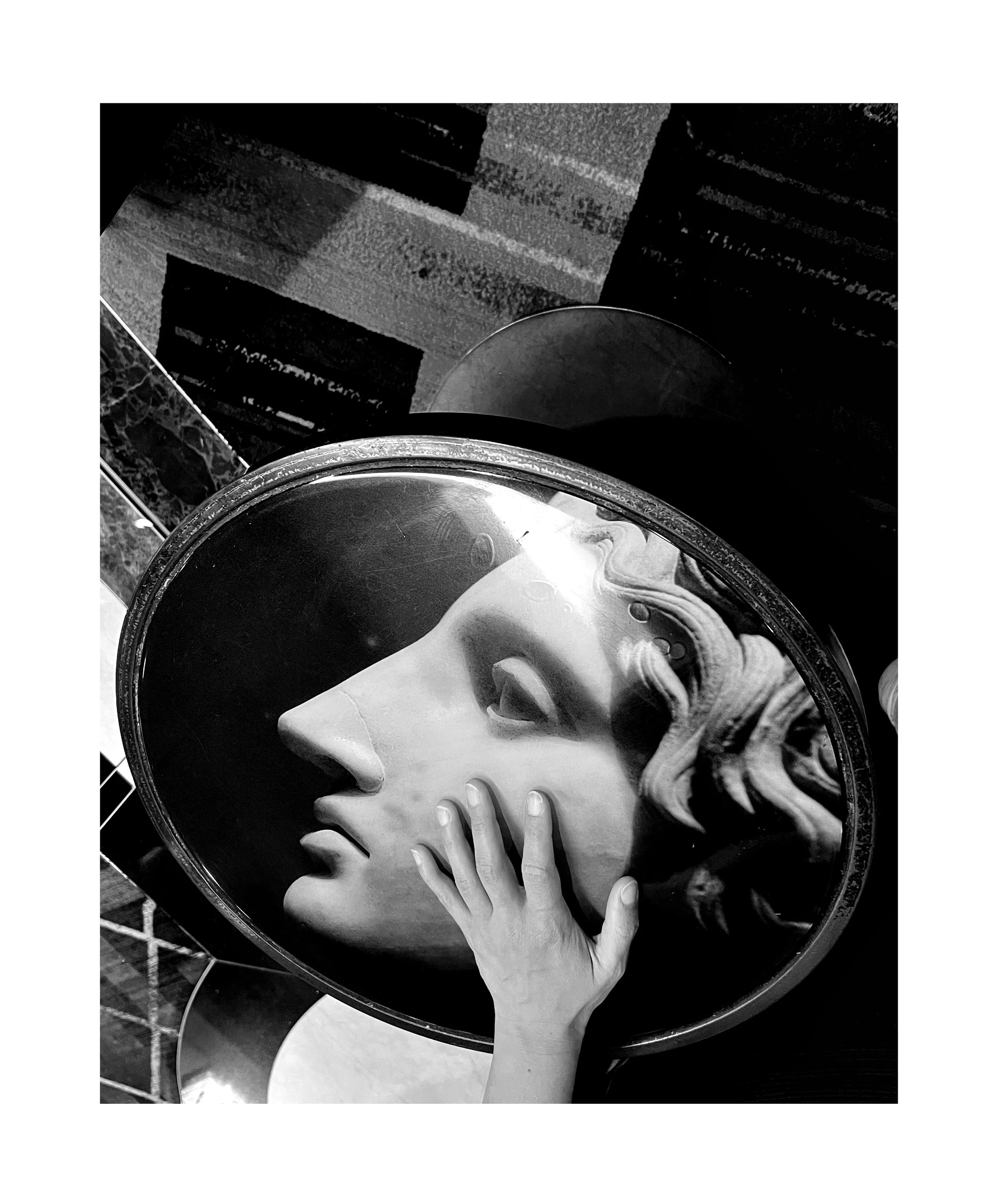 Une sculpture d'une tête de femme avec un visage expressif, vue à travers un miroir rond, avec une main humaine touchant la sculpture.