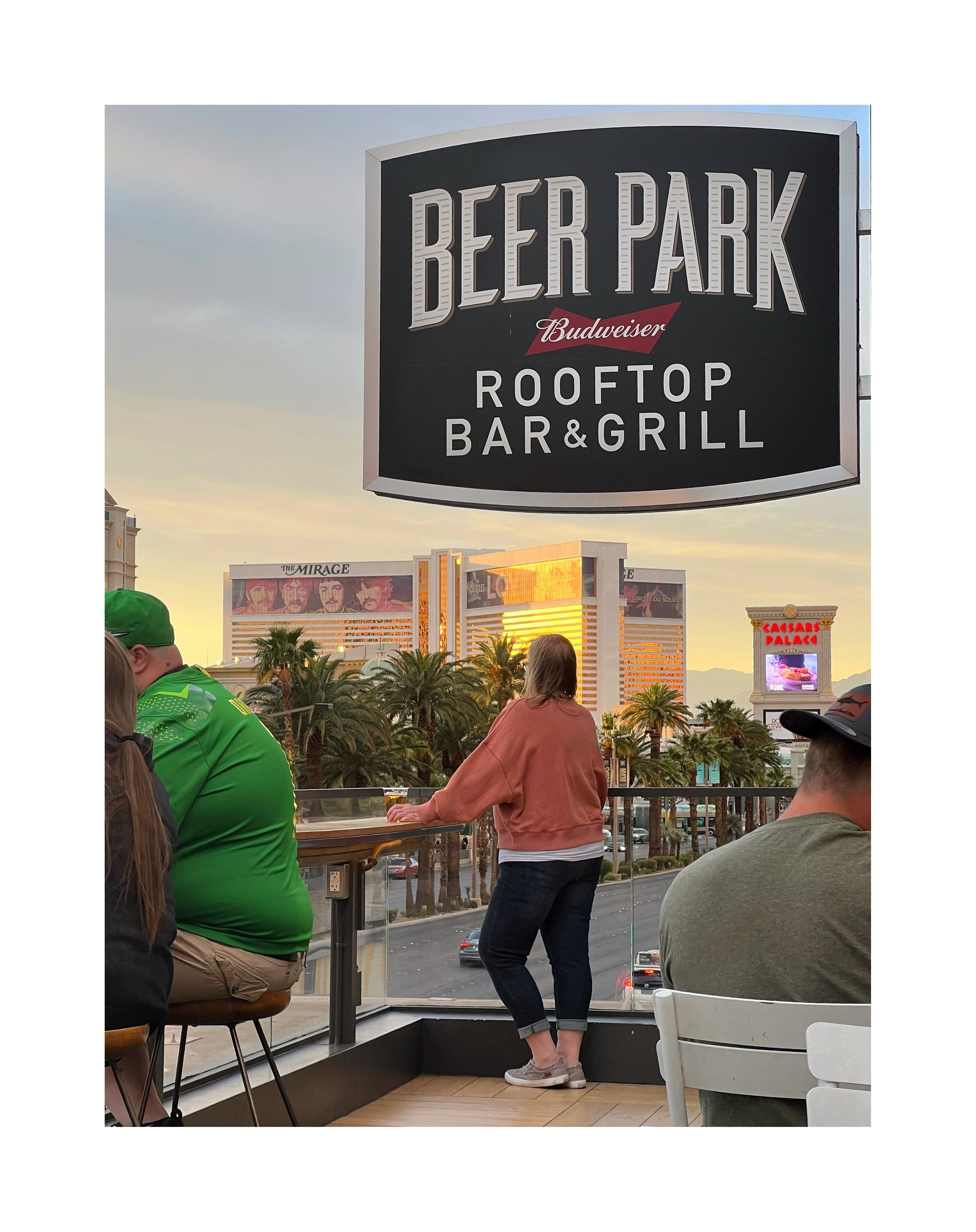 Photo d'une terrasse avec vue sur Las Vegas, montrant une femme debout près de la fenêtre, et des gens assis. Un panneau indique 'Beer Park Rooftop Bar & Grill'.