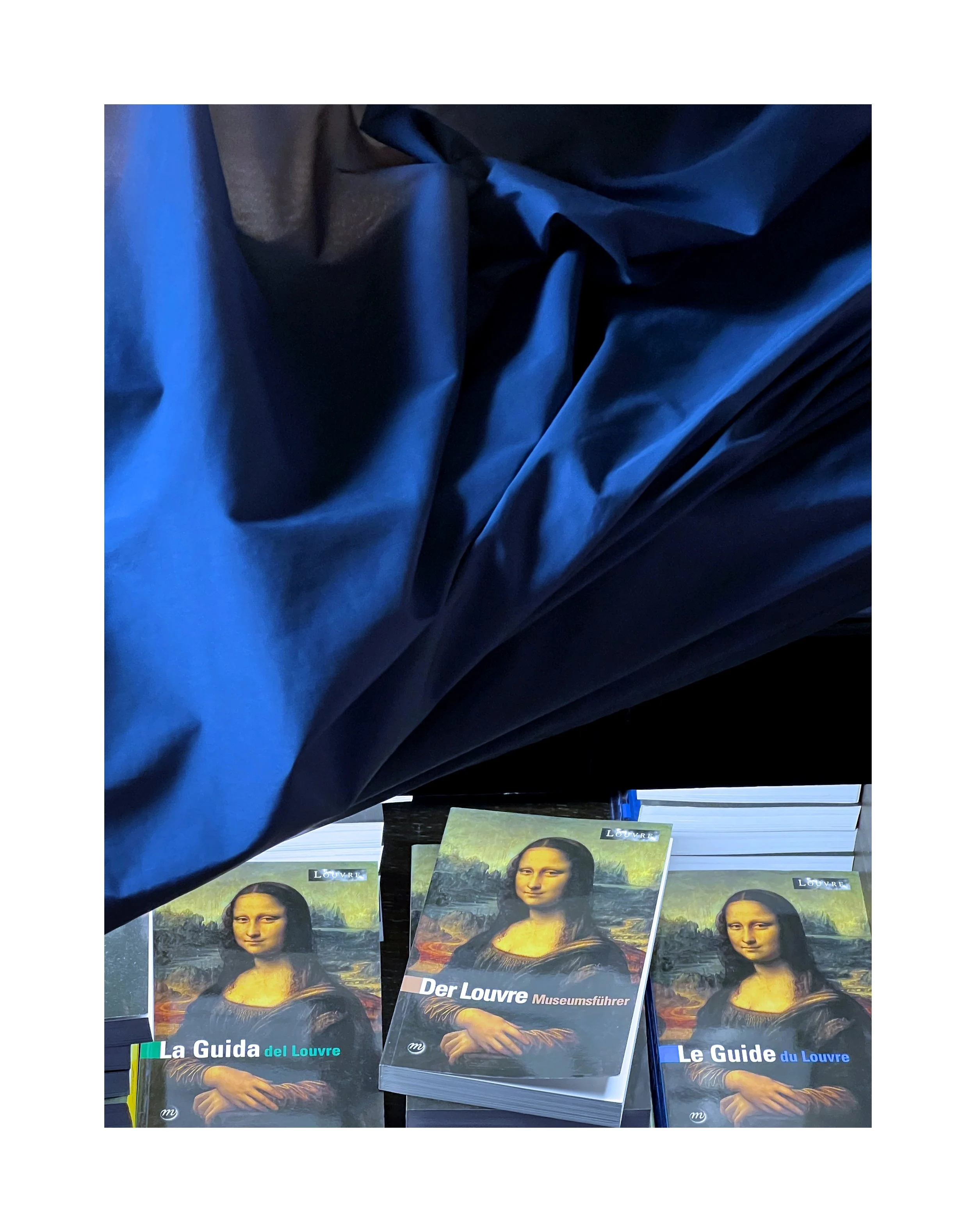 Des livres sur le tableau de La Joconde, un guide du Louvre en allemand, français et anglais, exposés sur une table, avec un tissu bleu en haut de l'image.