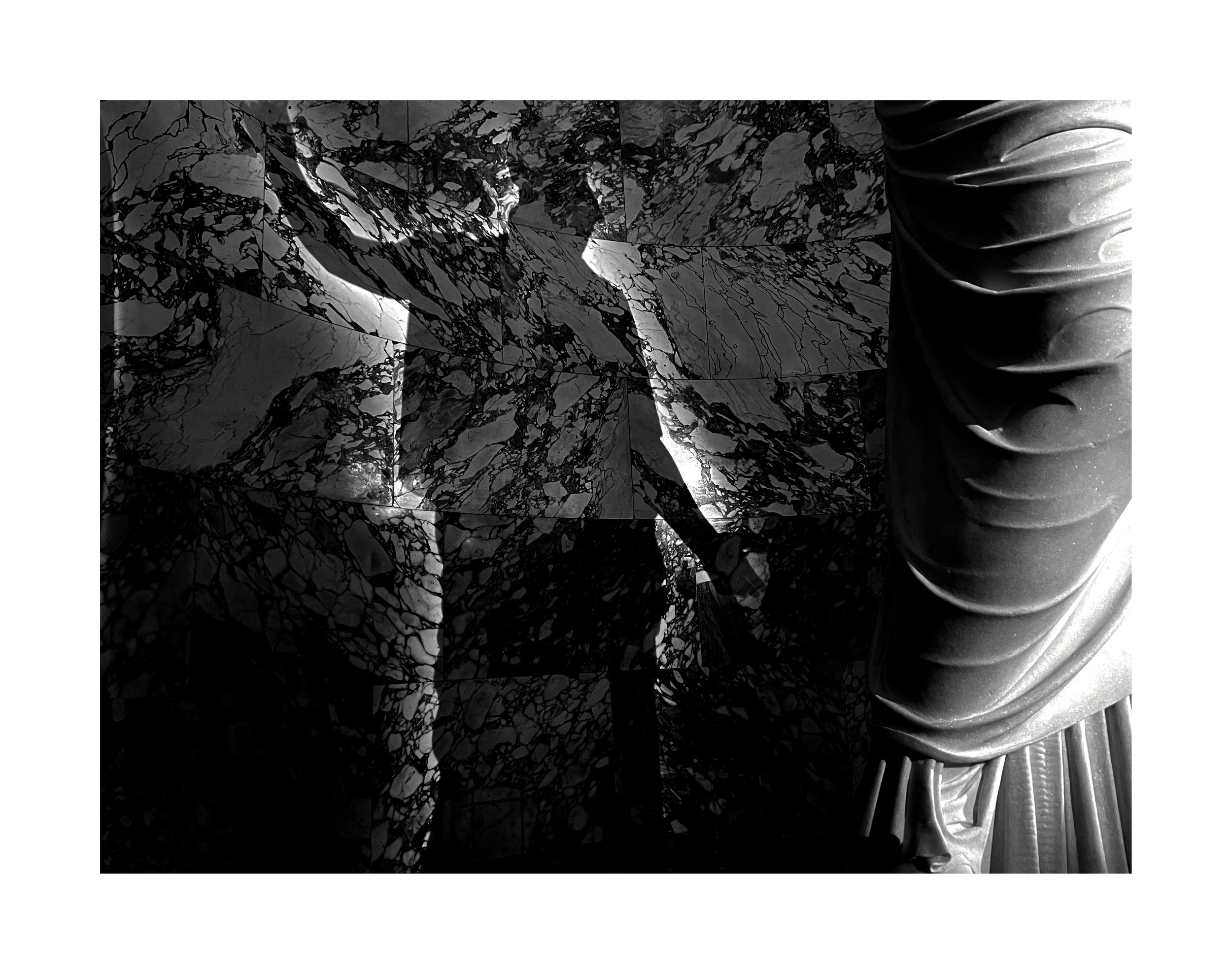 Une photo en noir et blanc montrant une sculpture en marbre avec un drapé ample à droite et un mur en marbre à motifs à gauche.