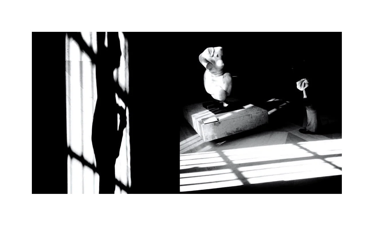 Une sculpture en forme de tête humaine, posée sur une caisse en bois, dans une pièce sombre avec de fortes ombres de fenêtres projetées sur le sol.