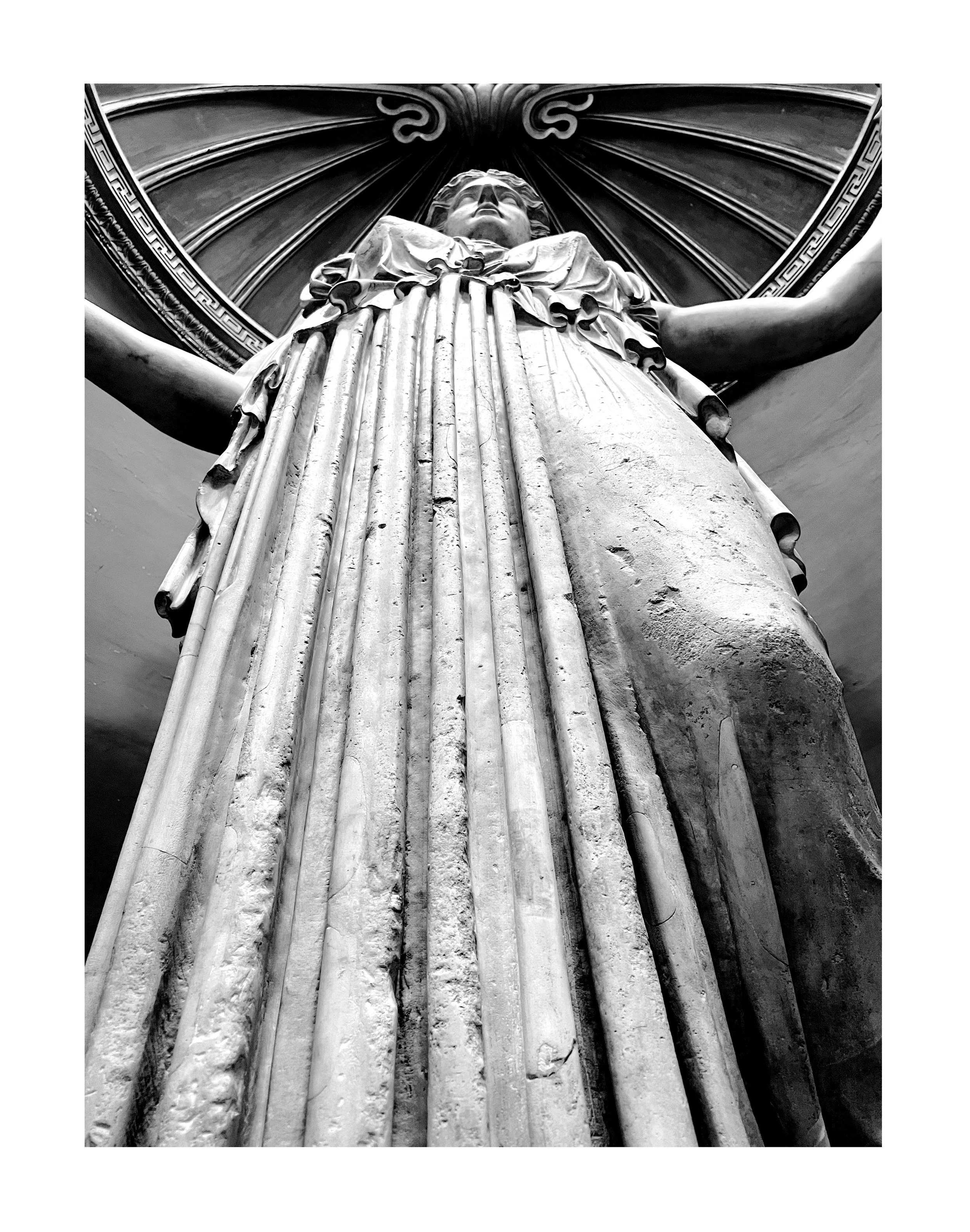 Photo en noir et blanc d'une statue recouverte d'une coiffure élaborée avec des bras étendus et une tête tournée vers le haut, placée dans un cadre architectural décoratif.