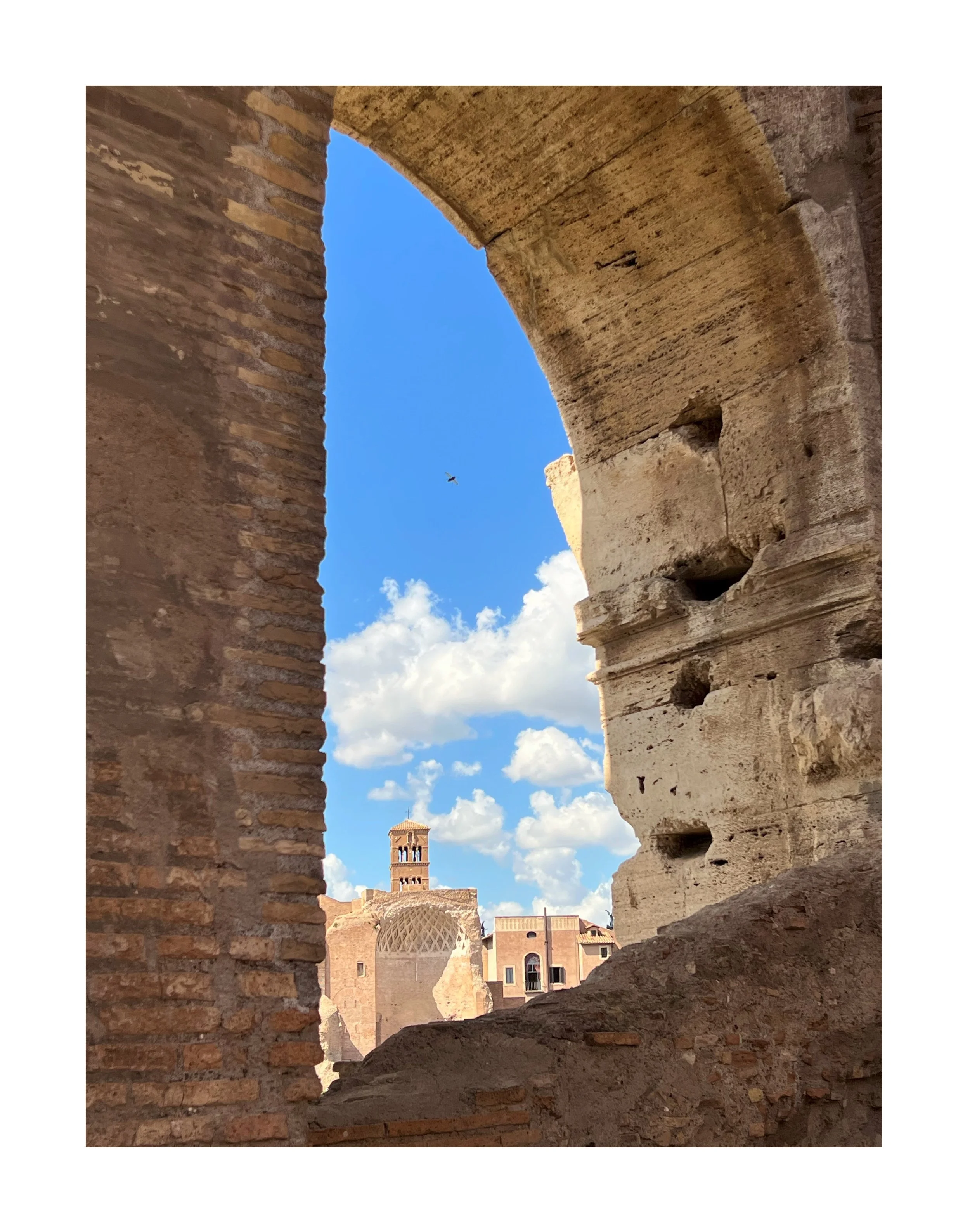 Vue à travers une ouverture dans un mur en ruines en direction d'une vieille ville avec un clocher, sous un ciel bleu avec quelques nuages.