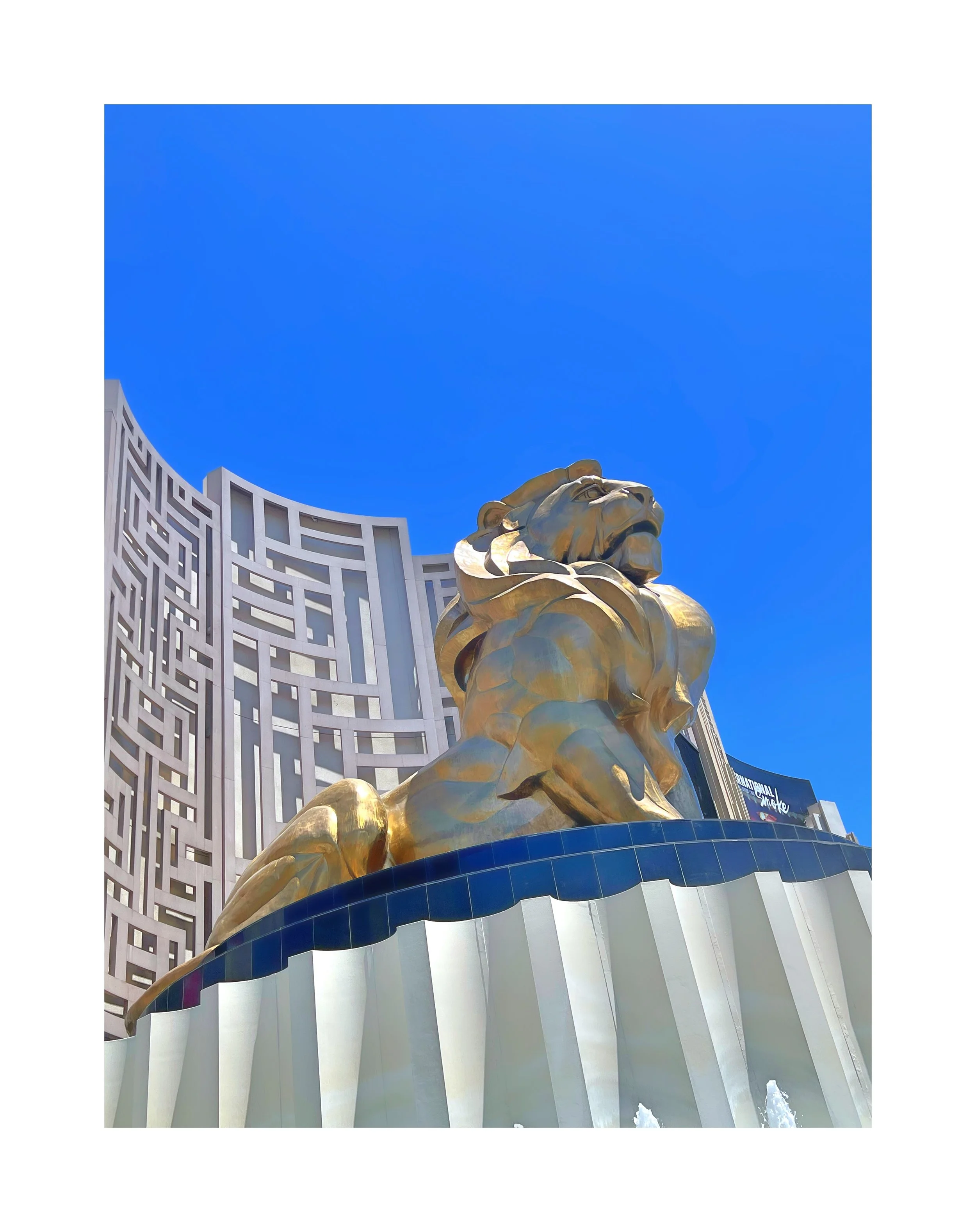 Une grande statue dorée d'un lion, en face d'un bâtiment avec une façade en treillis géométrique, sous un ciel bleu sans nuages.
