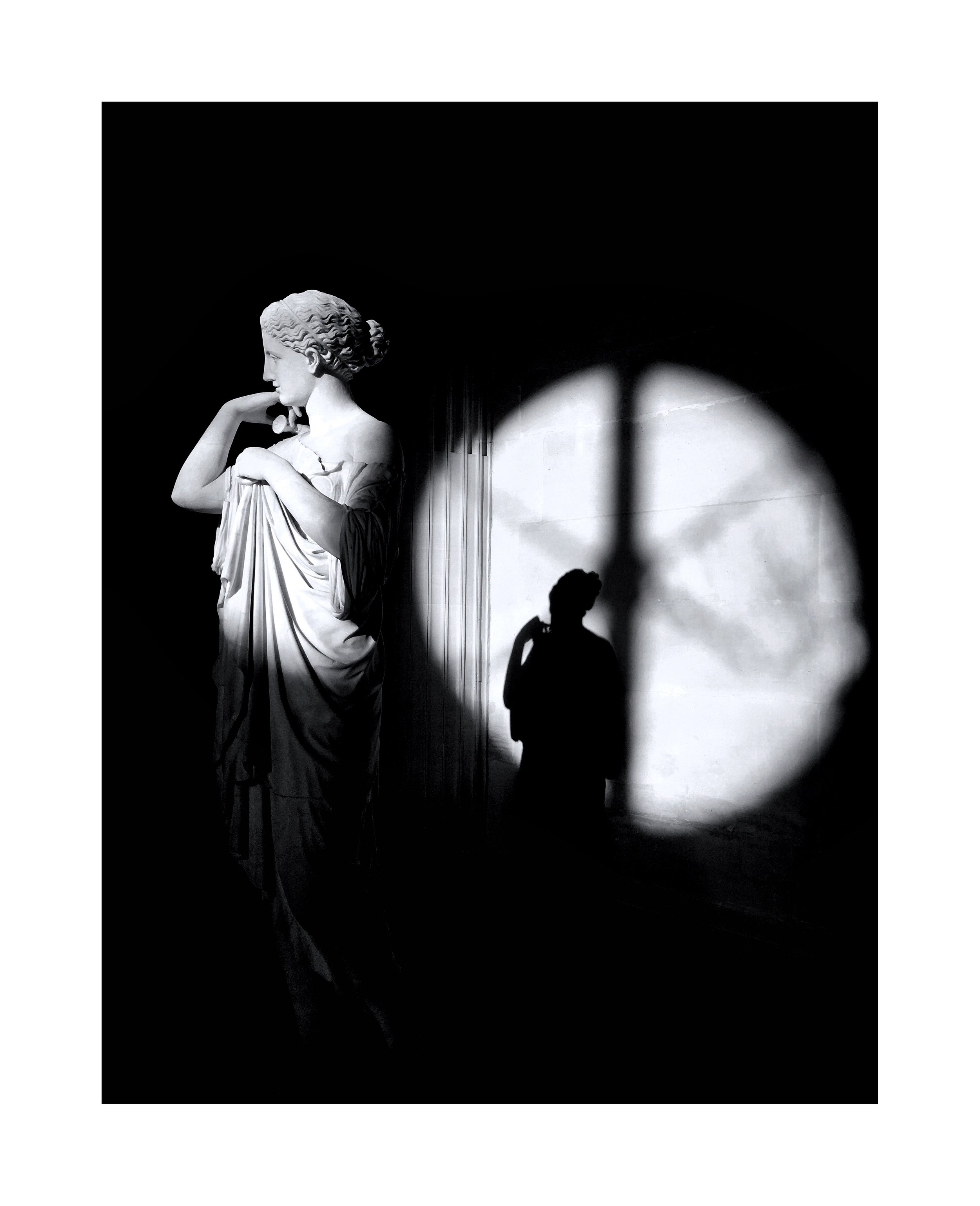 Une sculpture classique de femme en marbre avec ombre projetée sur un mur blanc, où une silhouette humaine est visible.