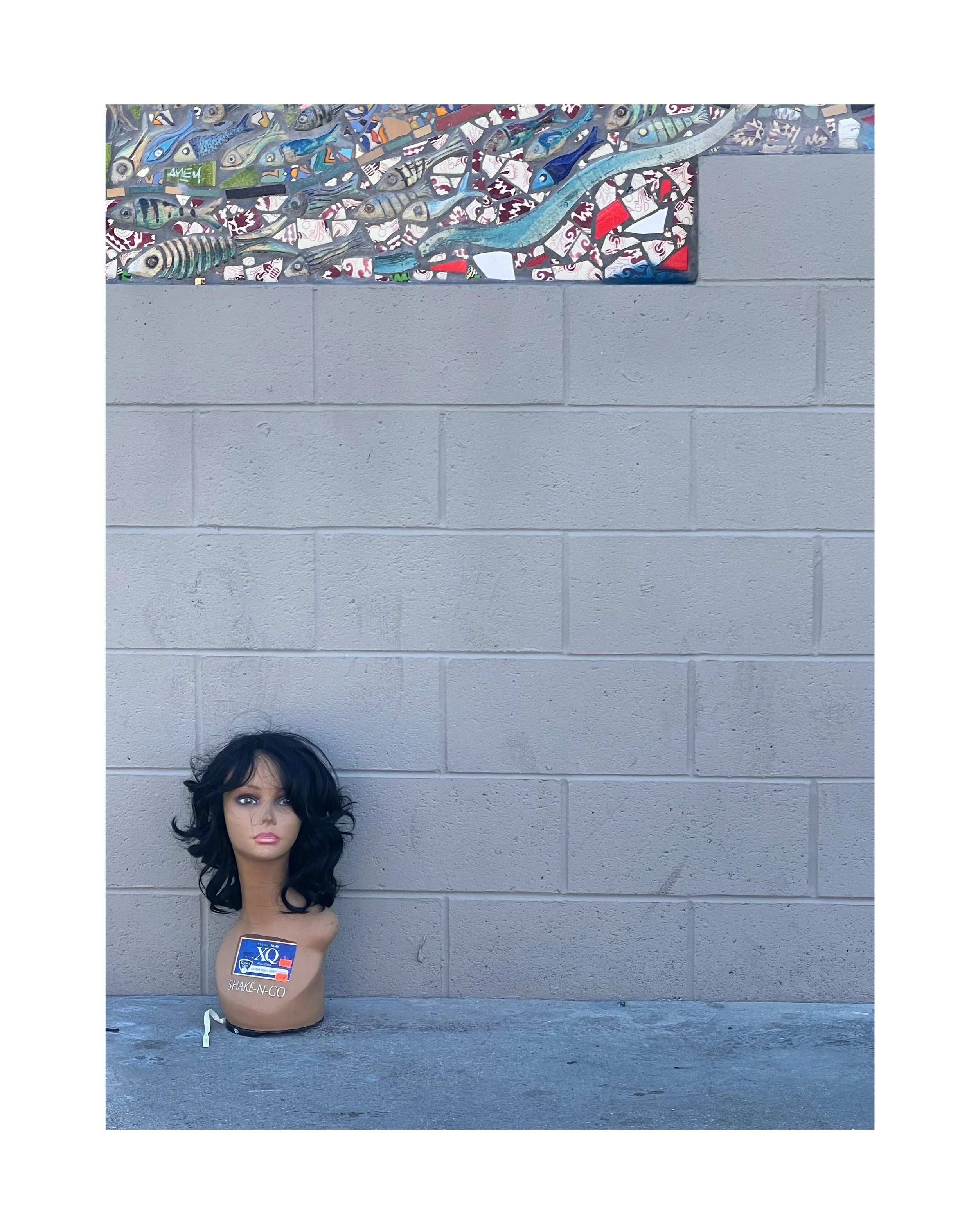 Poupée mannequin avec cheveux noirs bouclés et maquillage, placée sur une surface en pierre devant un mur en parpaings, avec une peinture murale colorée représentant des poissons en haut de l'image.