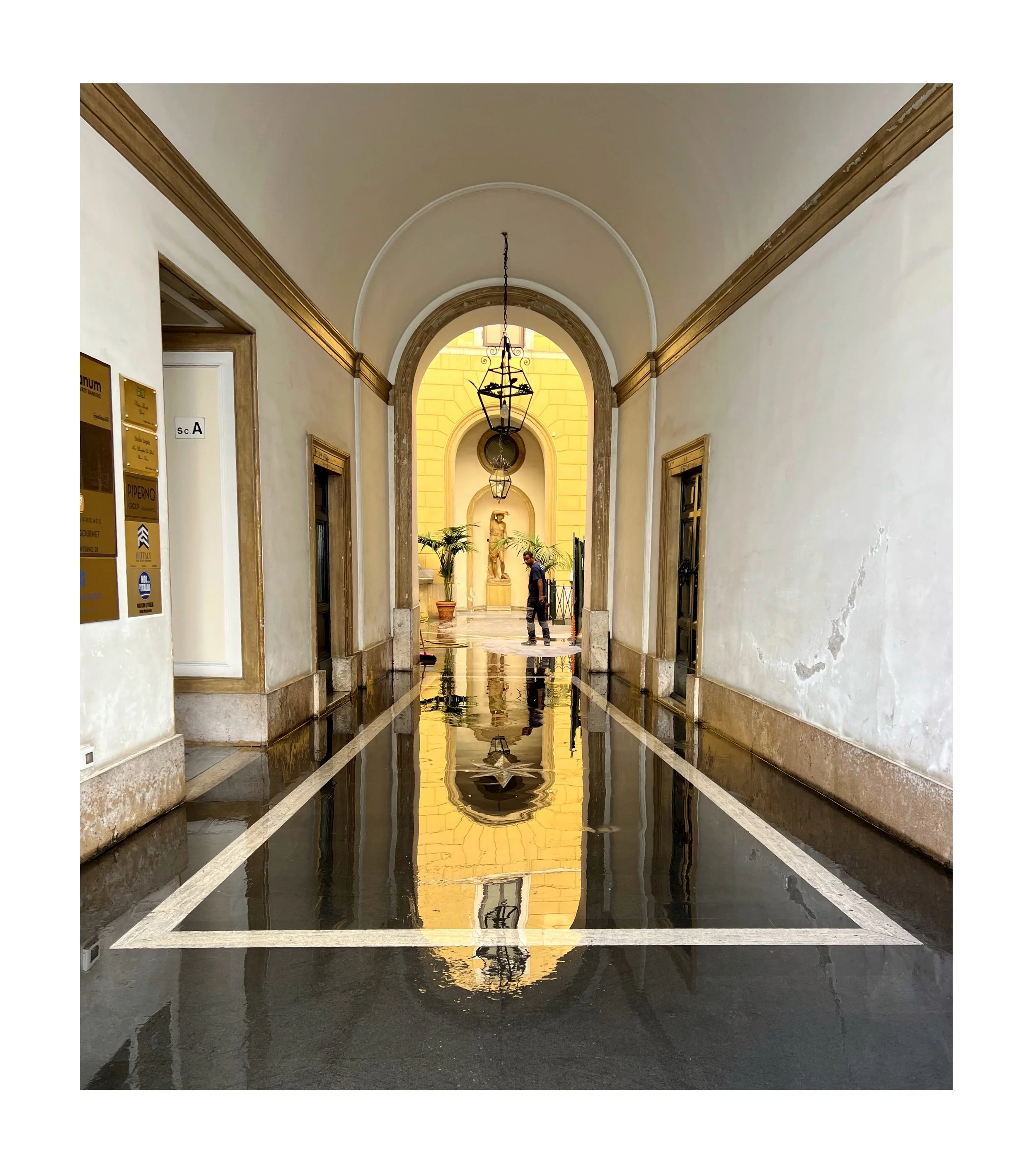 Intérieur d’un passage couvert avec un sol réfléchissant, des murs beige, des lampes suspendues et des portes. En arrière-plan, une statue et un homme regardant.