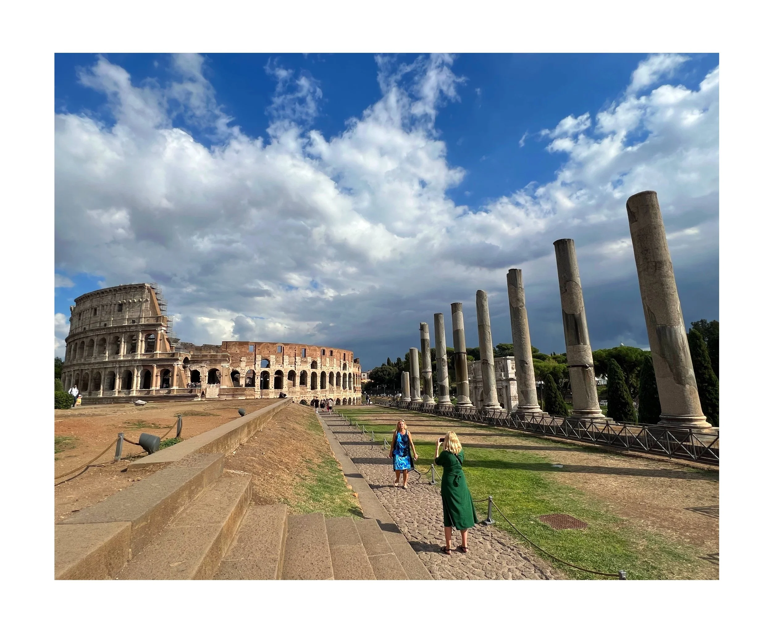 Le Colisée à Rome avec des colonnes antiques alignées sur la droite et un ciel nuageux, deux femmes en vêtements colorés se tenant sur un chemin pavé et prenant des photos, et un terrain en pente avec des marches en premier plan.