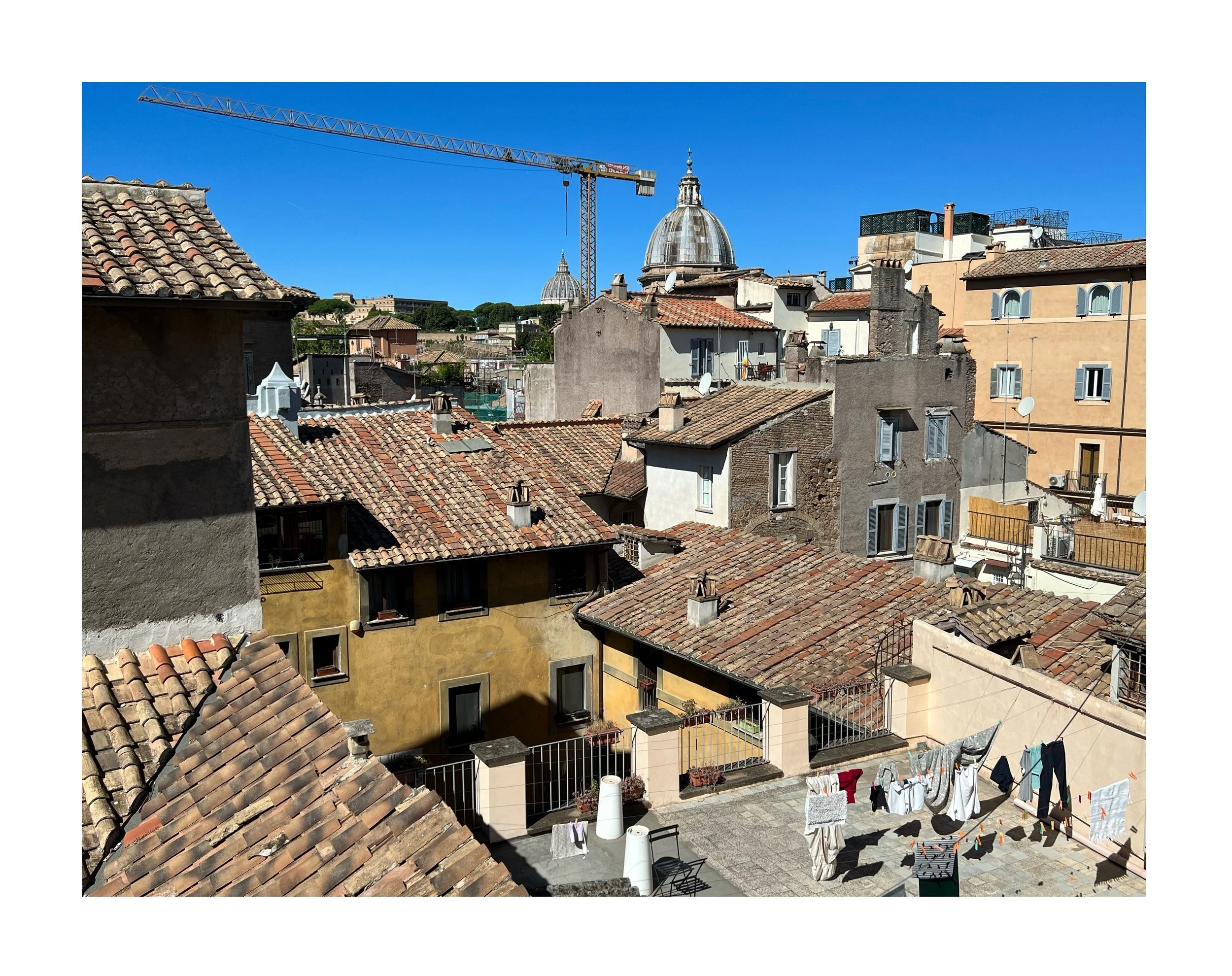 Vue d'un quartier résidentiel avec des toits en tuiles, un bâtiment avec un toit jaune, et des cordes à linge avec du linge suspendu. En arrière-plan, des bâtiments avec des dômes et une grue de construction, sous un ciel bleu clair.