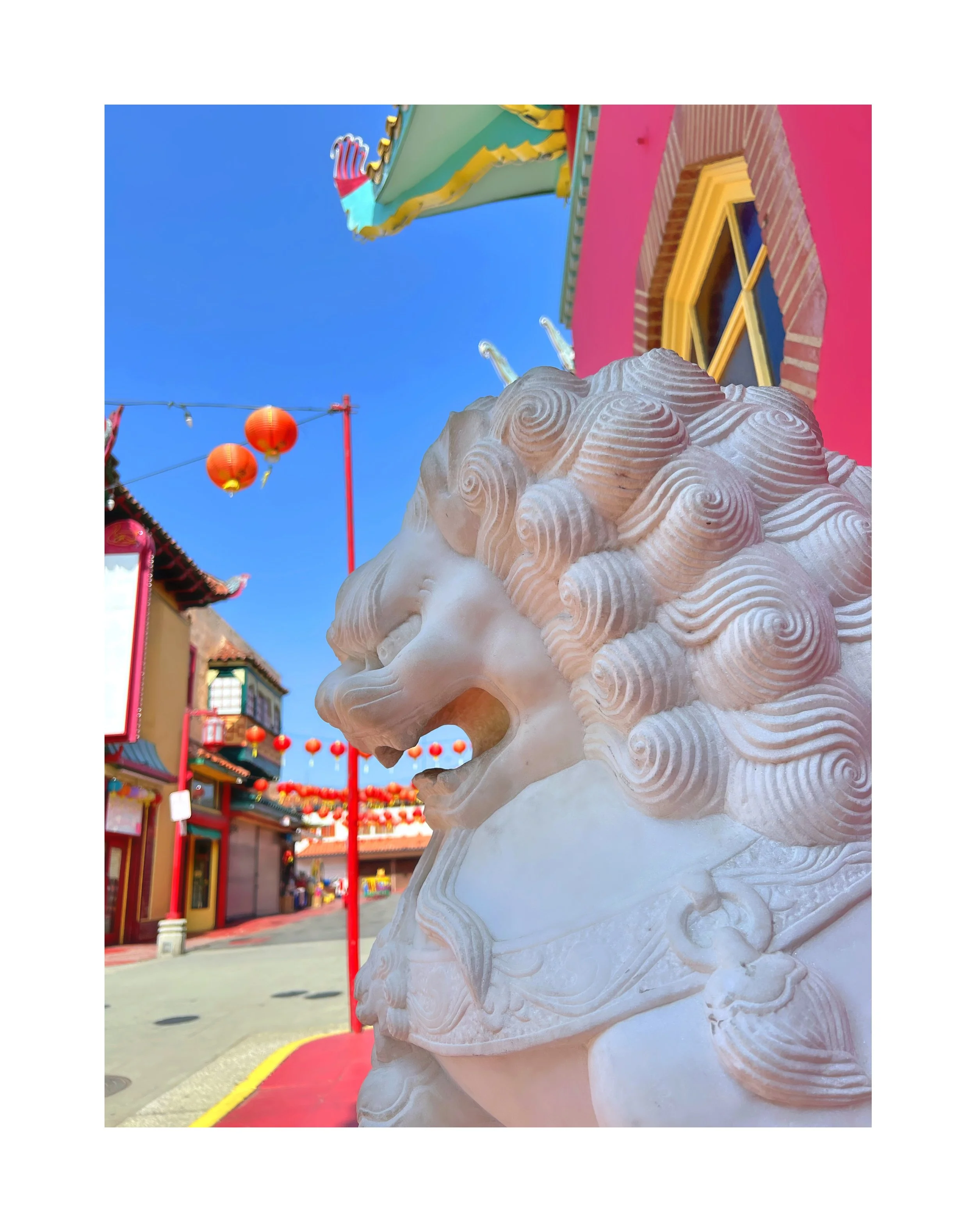 Statue de lion en pierre dans une rue décorée de lanternes rouges, bâtiments aux couleurs vives, ciel bleu clair.