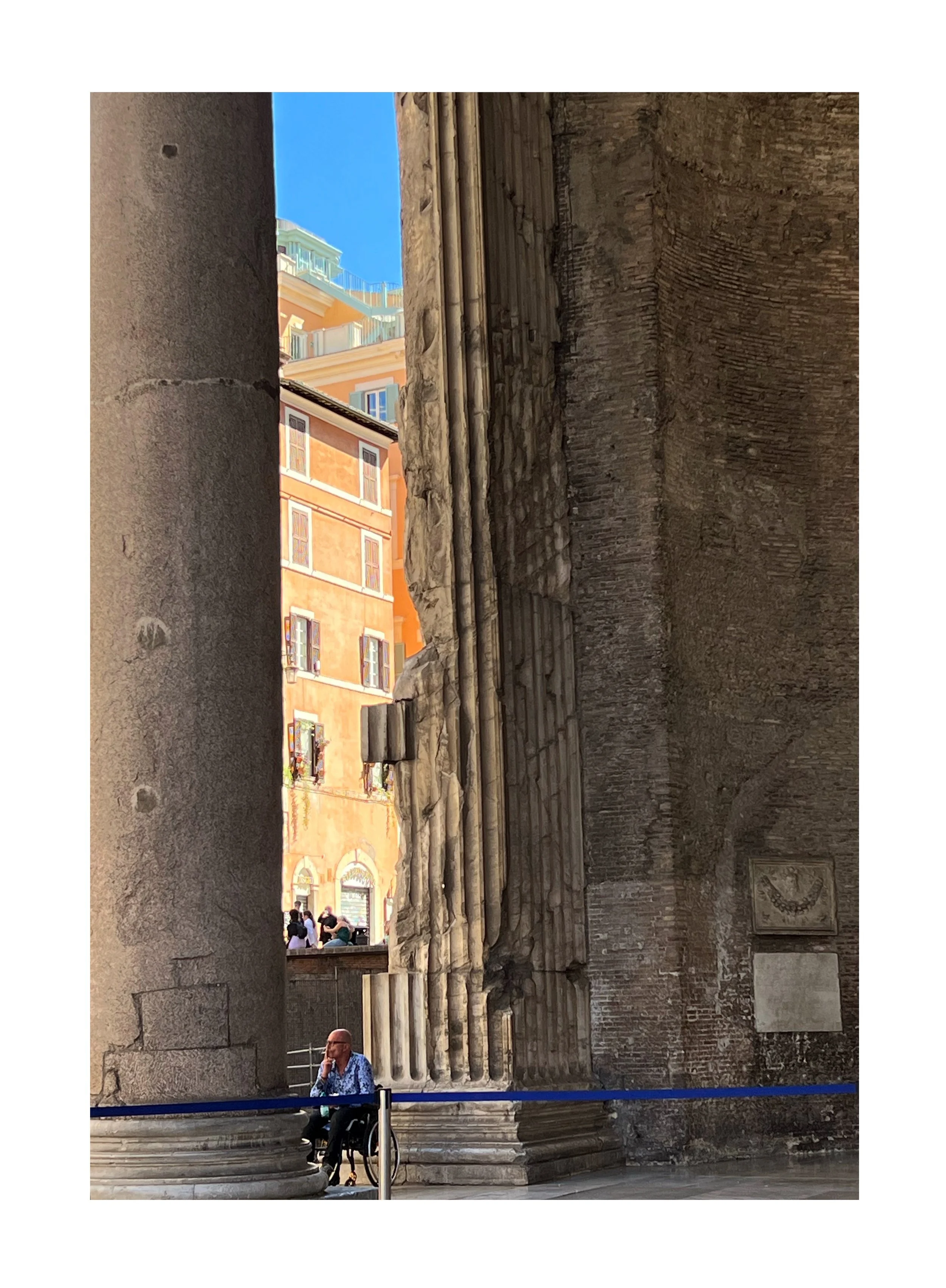 Photo d'une vieille structure romaine avec des colonnes, un homme assis en fauteuil roulant au premier plan, des bâtiments modernes colorés en arrière-plan, sous un ciel bleu clair.