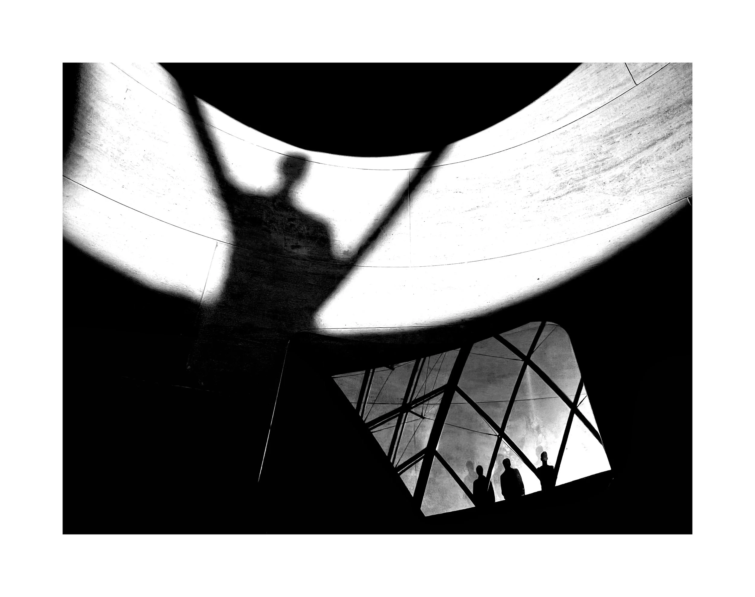 Silhouettes de trois personnes regardant par une grande fenêtre en verre, avec une ombre d'une figure humaine projetée sur un mur intérieur au-dessus, dans un bâtiment moderne avec des formes architecturales géométriques.