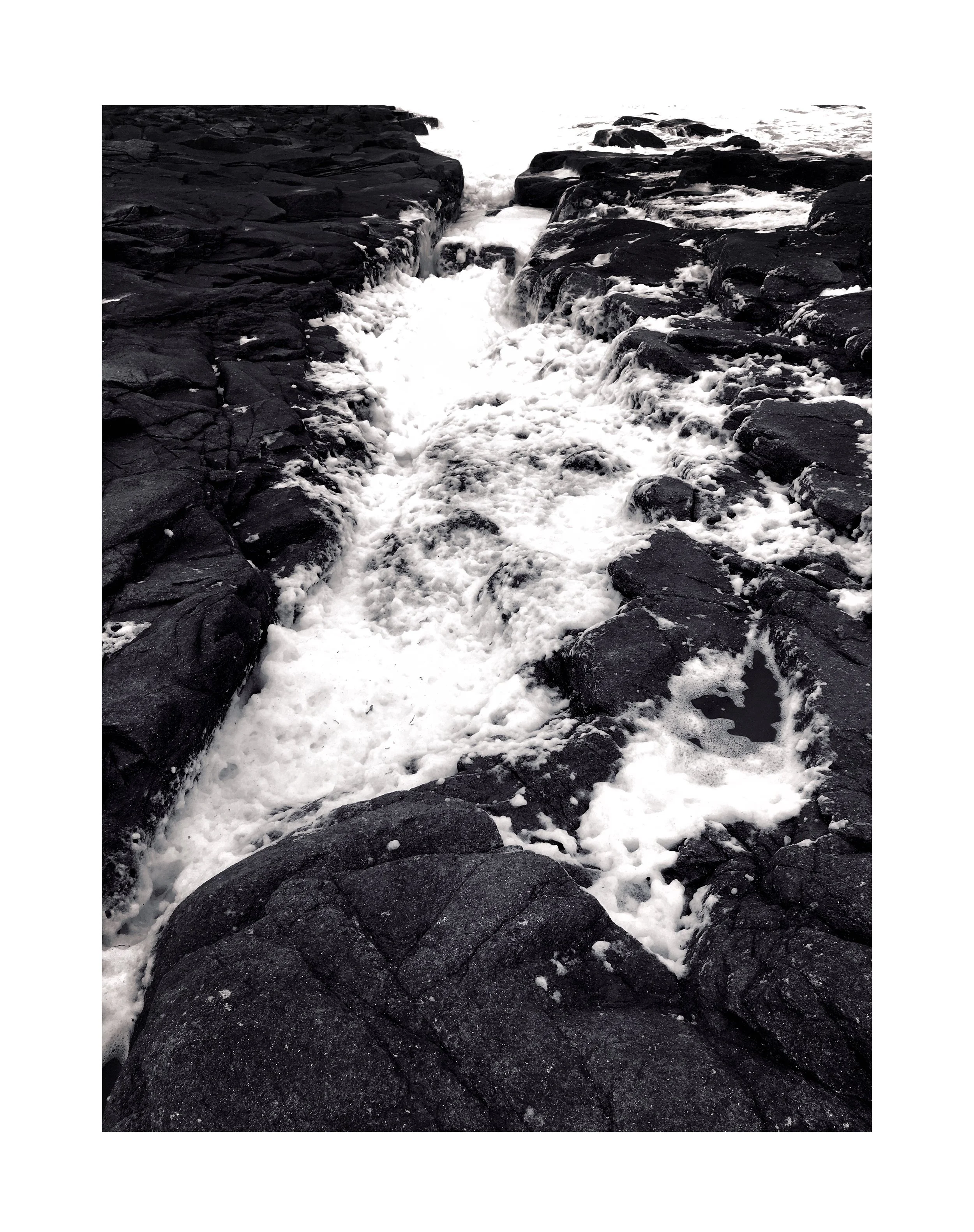 Vague d'eau qui coule sur des rochers noirs, créant des éclaboussures et de la mousse blanche.