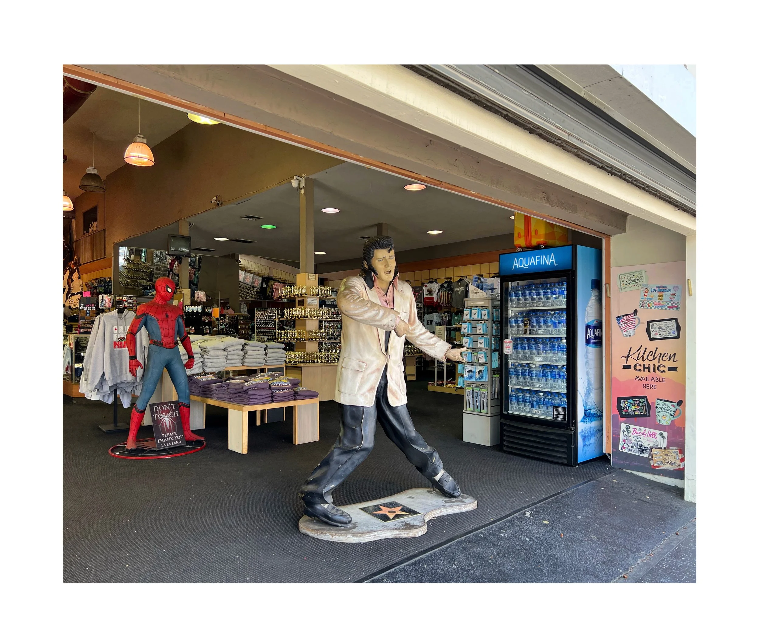 Statue de Elvis Presley à l'entrée d'une boutique, avec une figurine de Spider-Man à côté, et un réfrigérateur d'eau en vente à proximité.