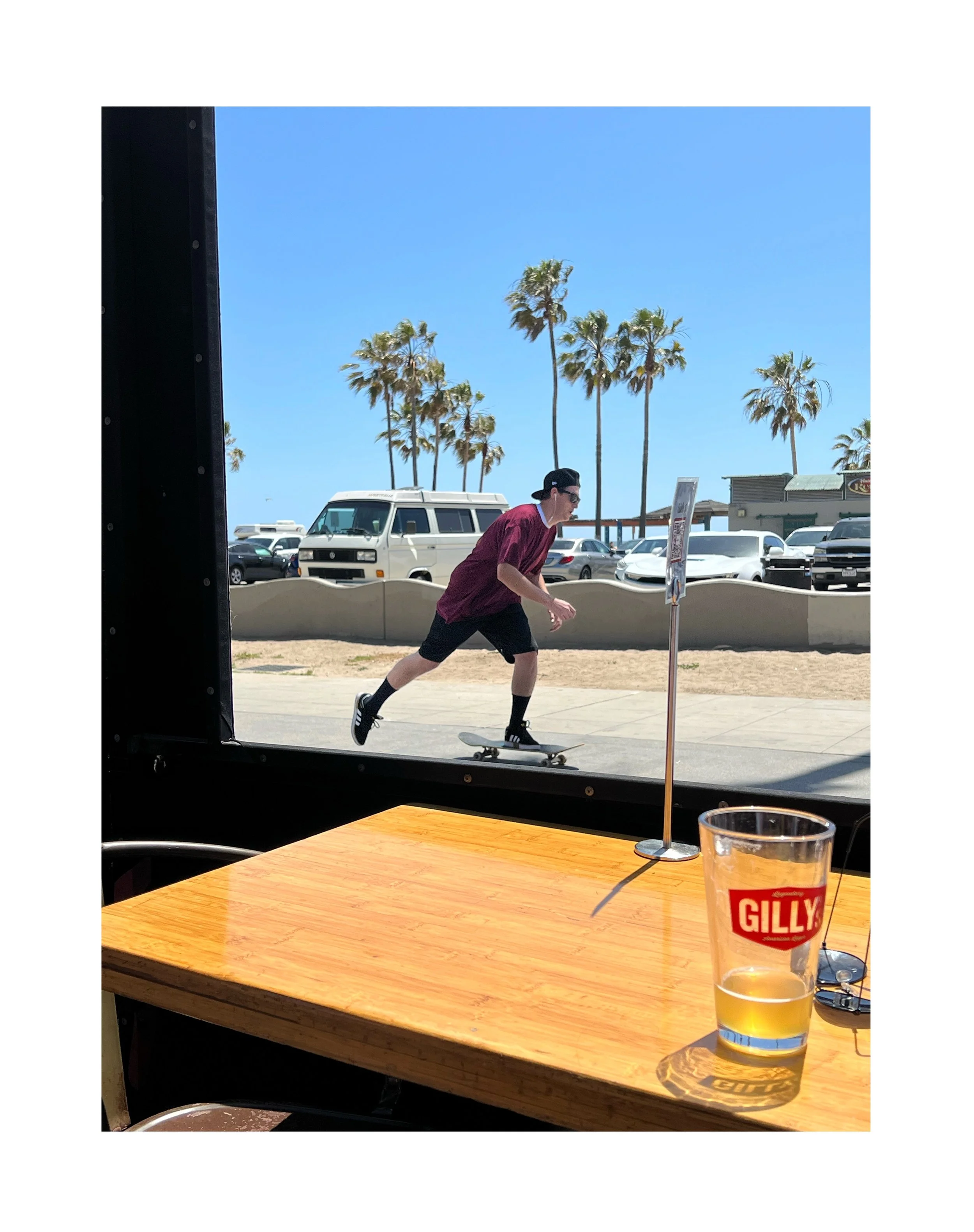 Jeune homme en skateboard à côté de la fenêtre d'un restaurant avec un verre de bière Gilly sur la table, vue sur des palmiers, des voitures et un ciel bleu.