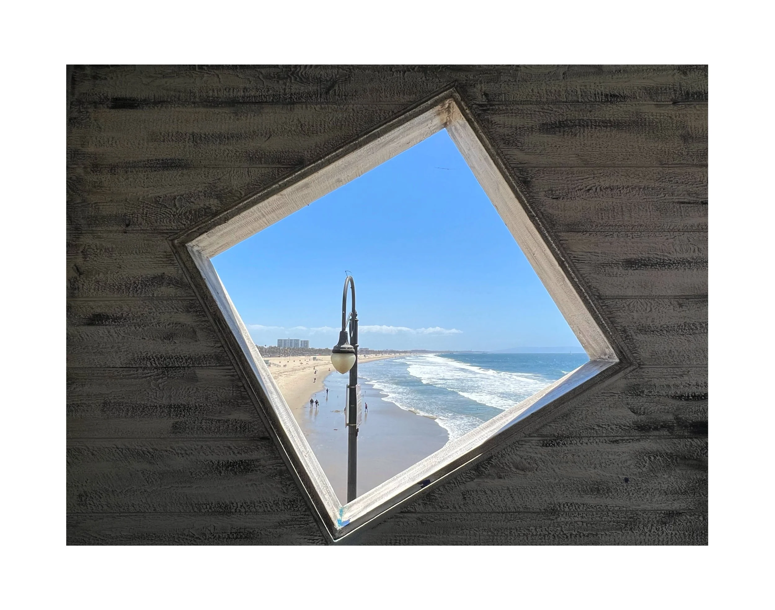 Une vue de la plage avec le sable, la mer et le ciel bleu, vue à travers une fenêtre en forme de diamant sur une cabane en bois.