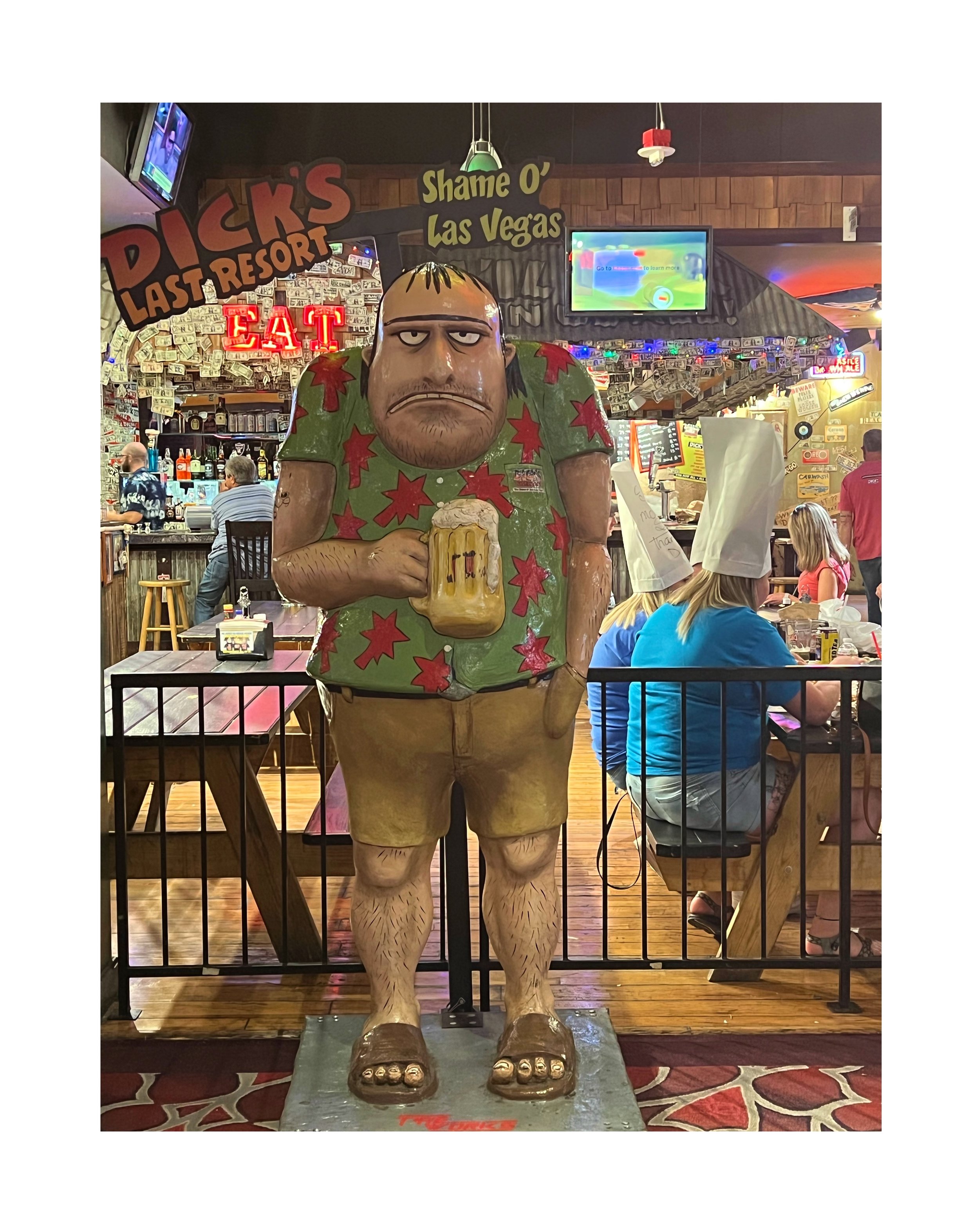 Statue de cartoon d'un homme gros avec une bière à la main dans un bar dîner, décorations et clients en arrière-plan.