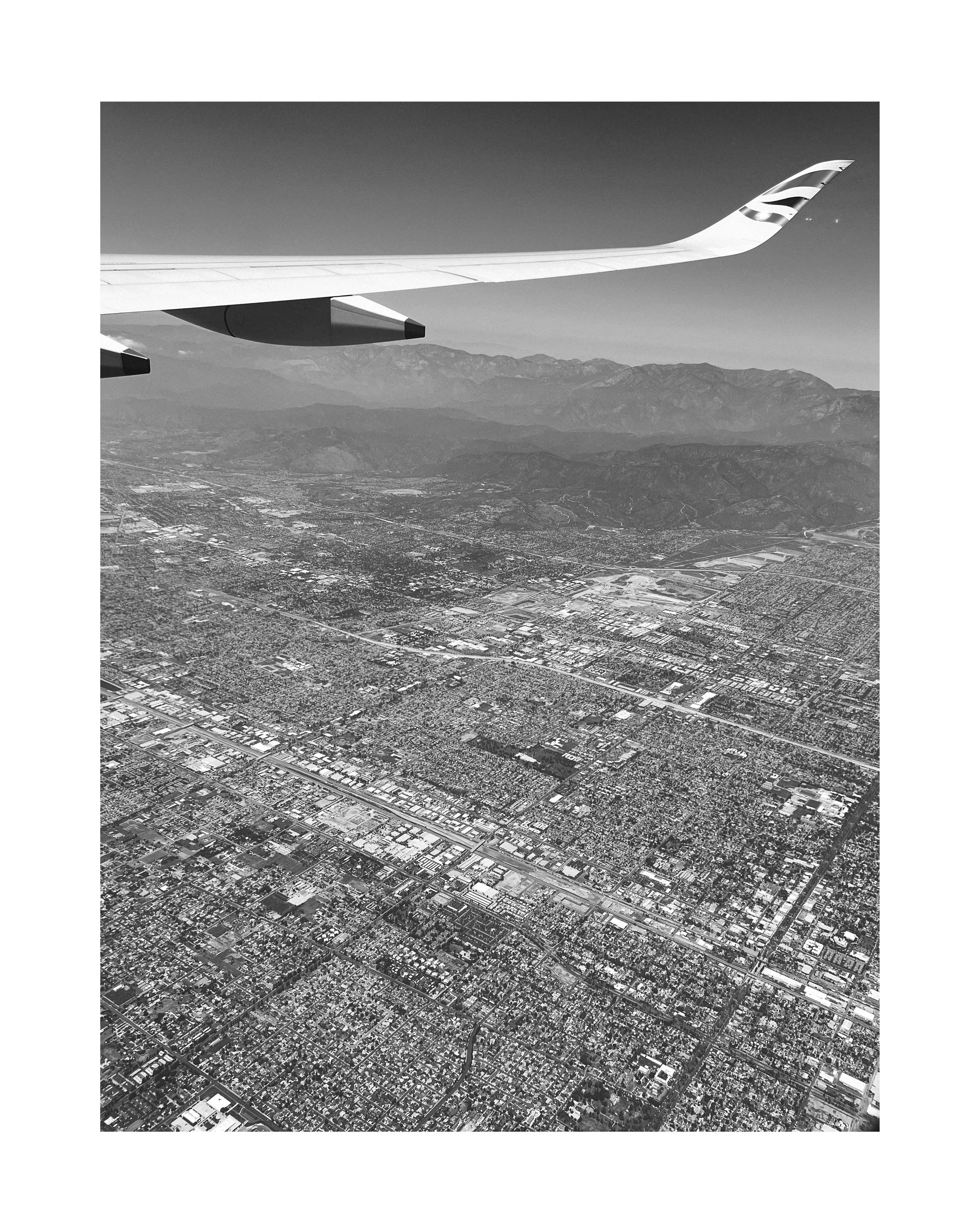 Vue aérienne d'une ville vue depuis un avion en vol, avec un paysage de montagnes à l'horizon, en noir et blanc.