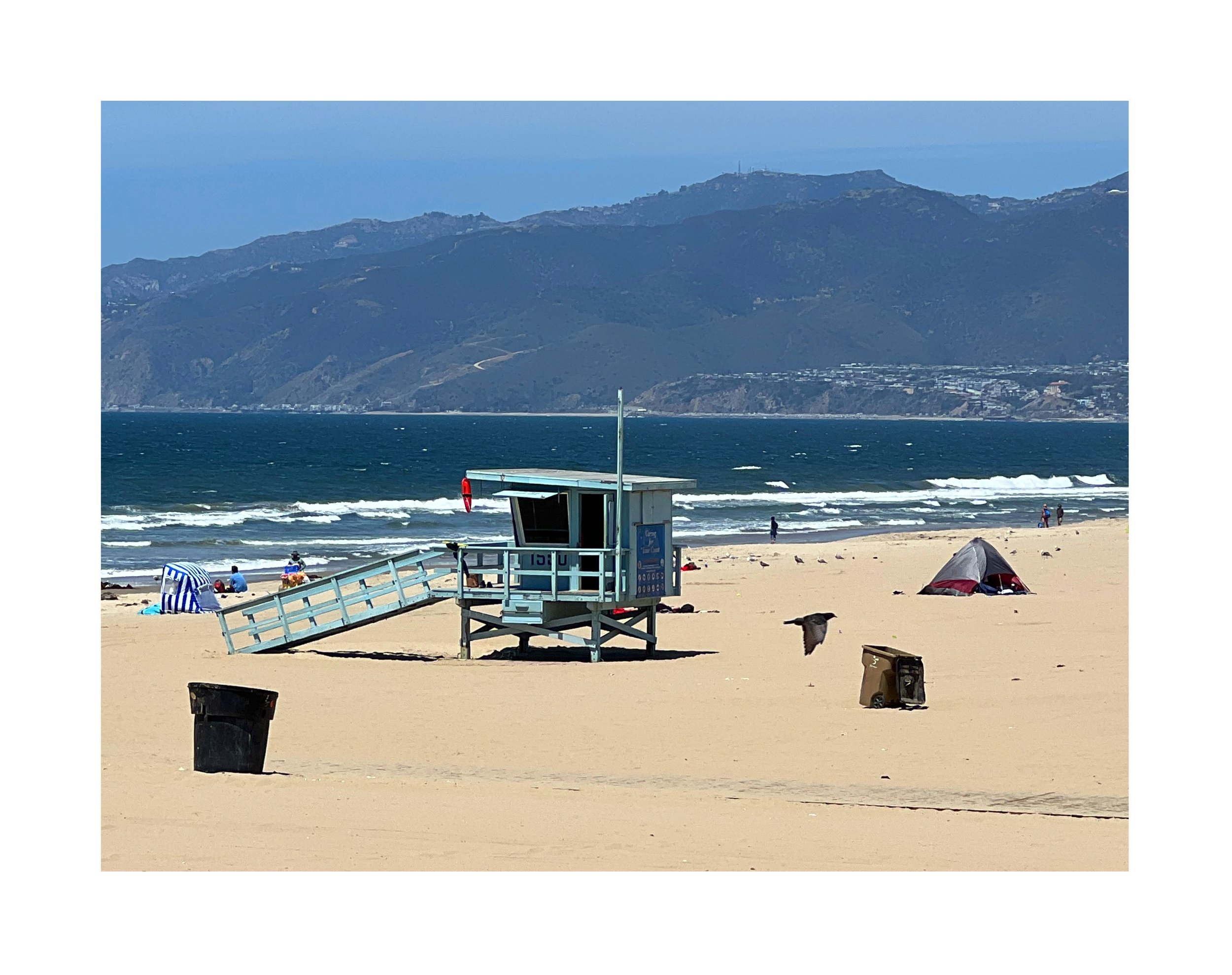 Plage avec des parasols, un poste de sauveteur, des personnes et un chien, avec des montagnes en arrière-plan et le ciel bleu.