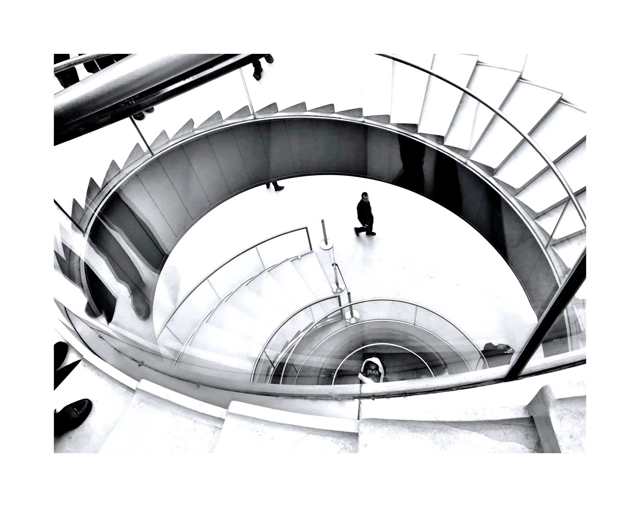 Vue d'en haut d'une cage d'escalier en spirale avec des marches en métal et un sol blanc, deux personnes descendent ou montent les escaliers, vue en noir et blanc.