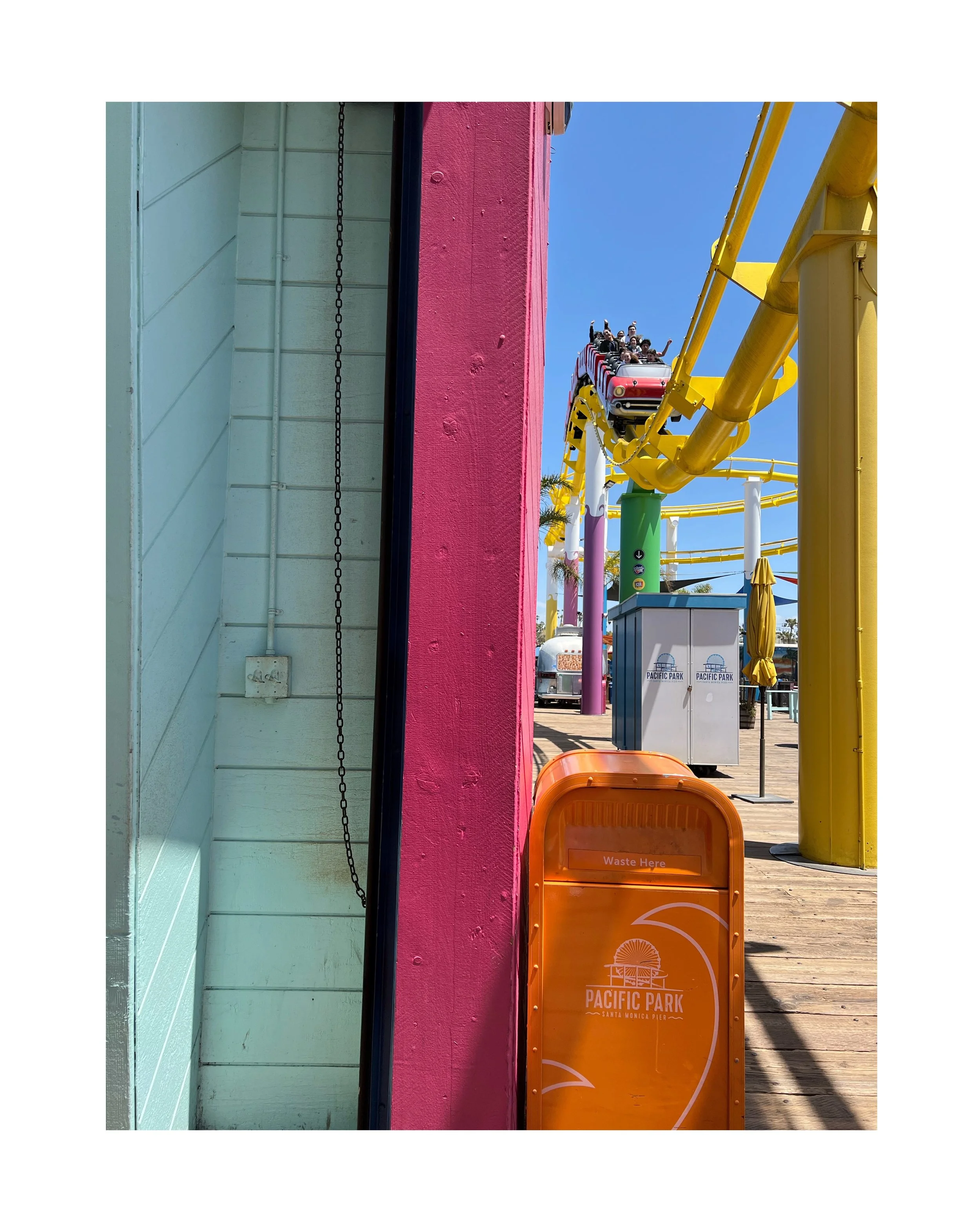Une corbeille à déchets orange avec écrit 'Waste Here' et logo du parc d'attractions Pacific Park Santa Monica, avec à l'arrière un pont roulant jaune et une file d'attente pour les montagnes russes.