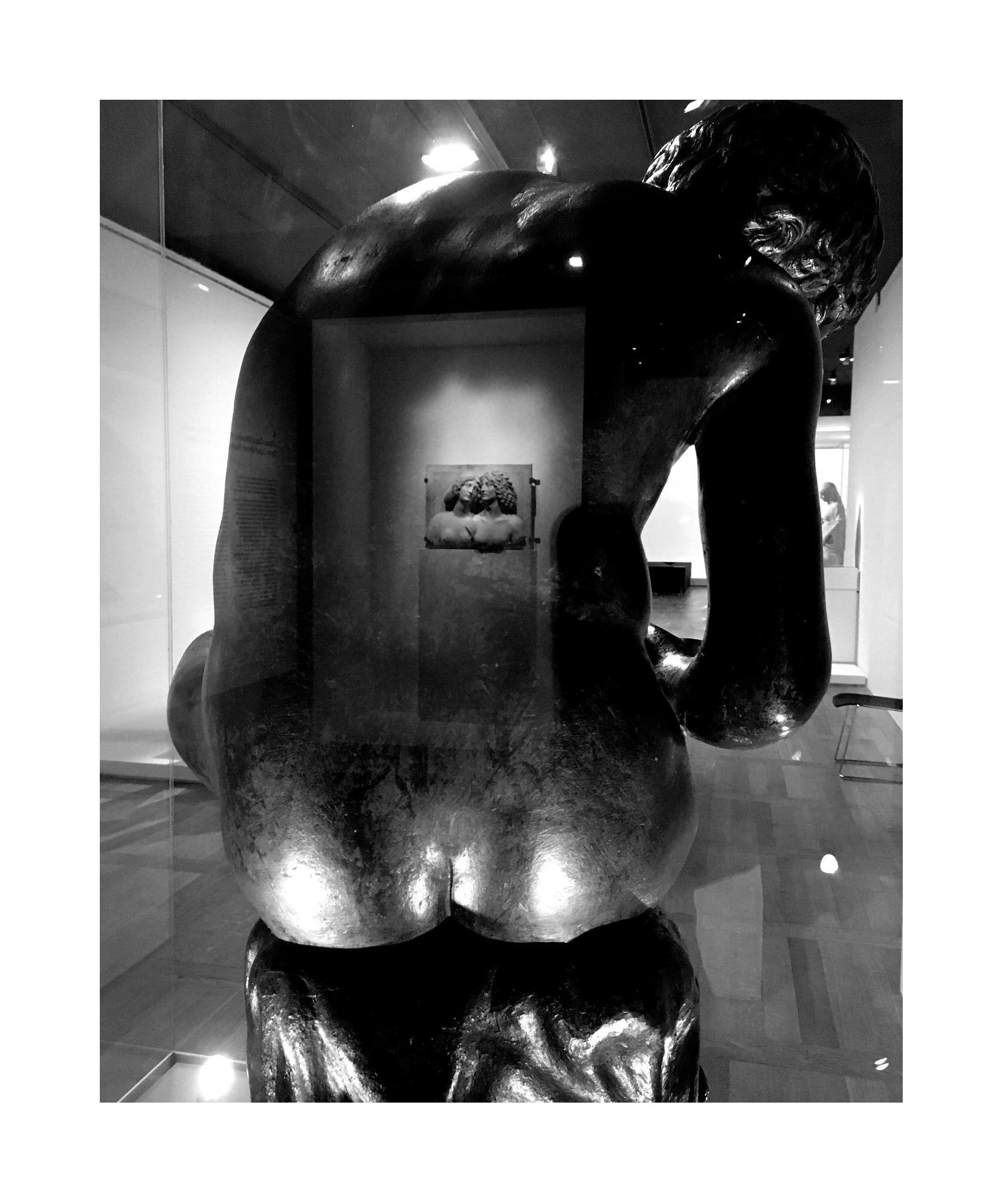 Sculpture en bronze représentant un homme penché, vue de dos, dans un musée, avec une peinture en arrière-plan.