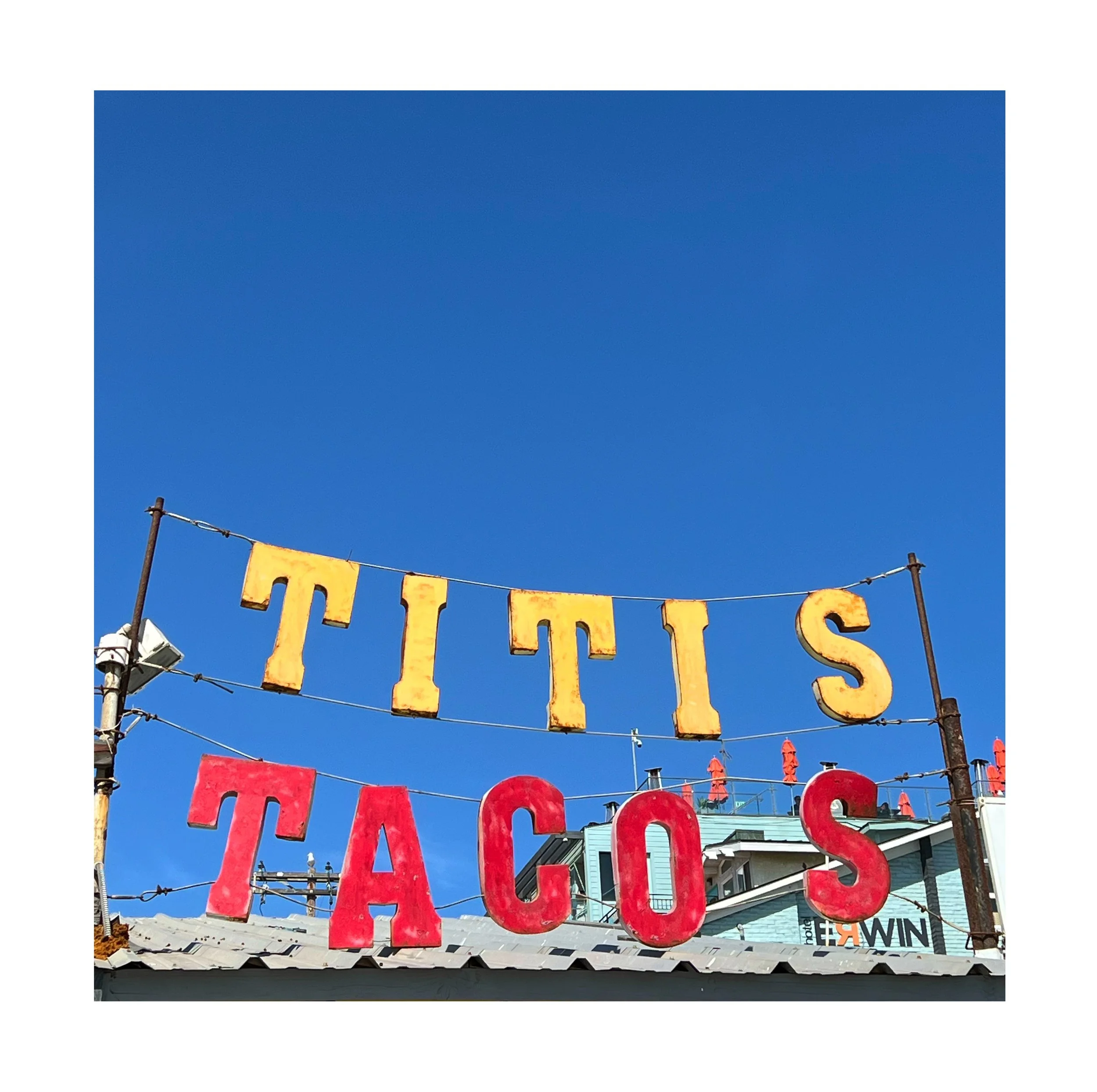 Une enseigne avec le texte « Titis Tacos » en lettres rouges et jaunes, suspendue au-dessus d'un bâtiment avec un ciel bleu clair en arrière-plan.