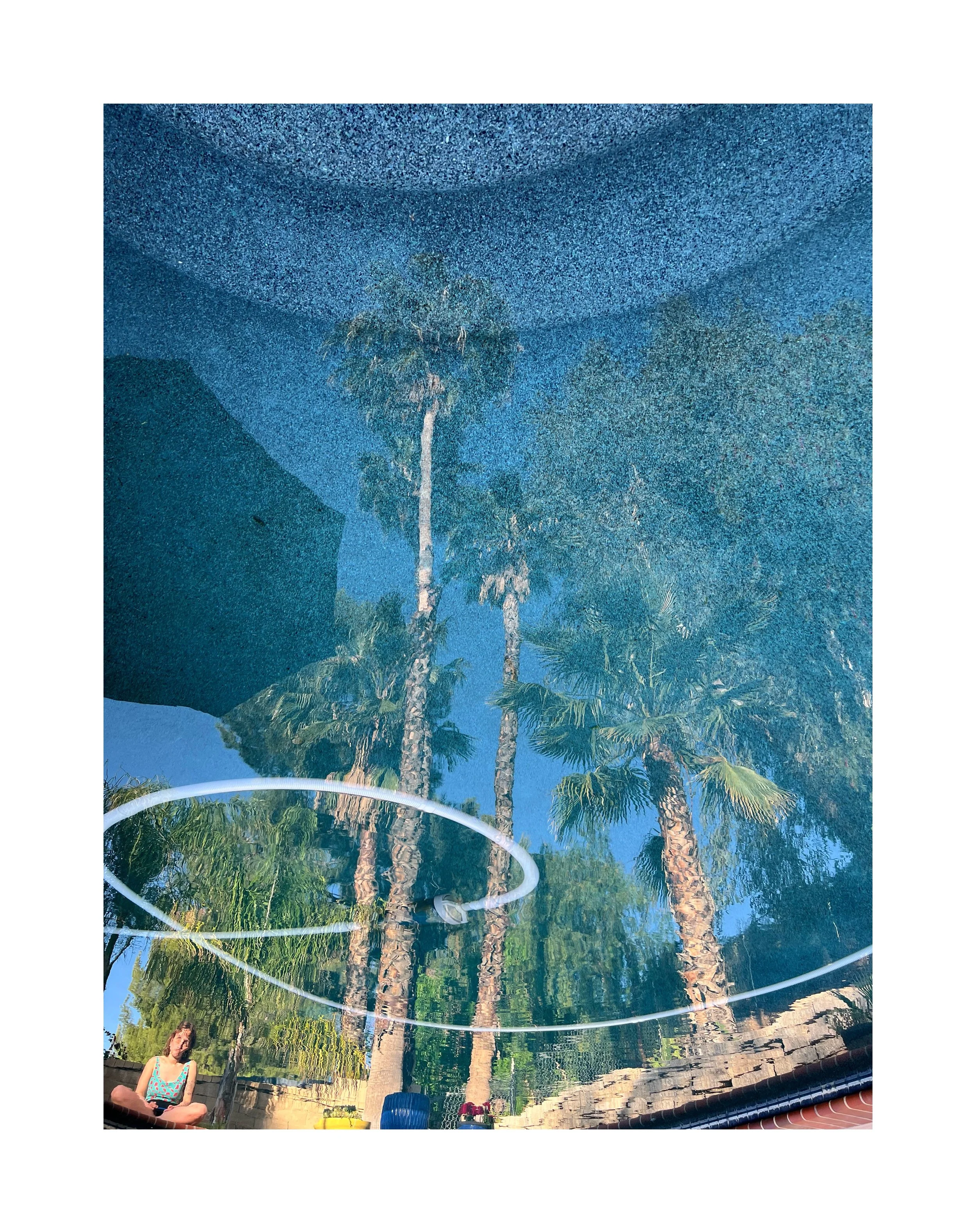 Reflet de palmiers et d'un ciel bleu dans une piscine, avec une femme assise au bord de la piscine dessous.