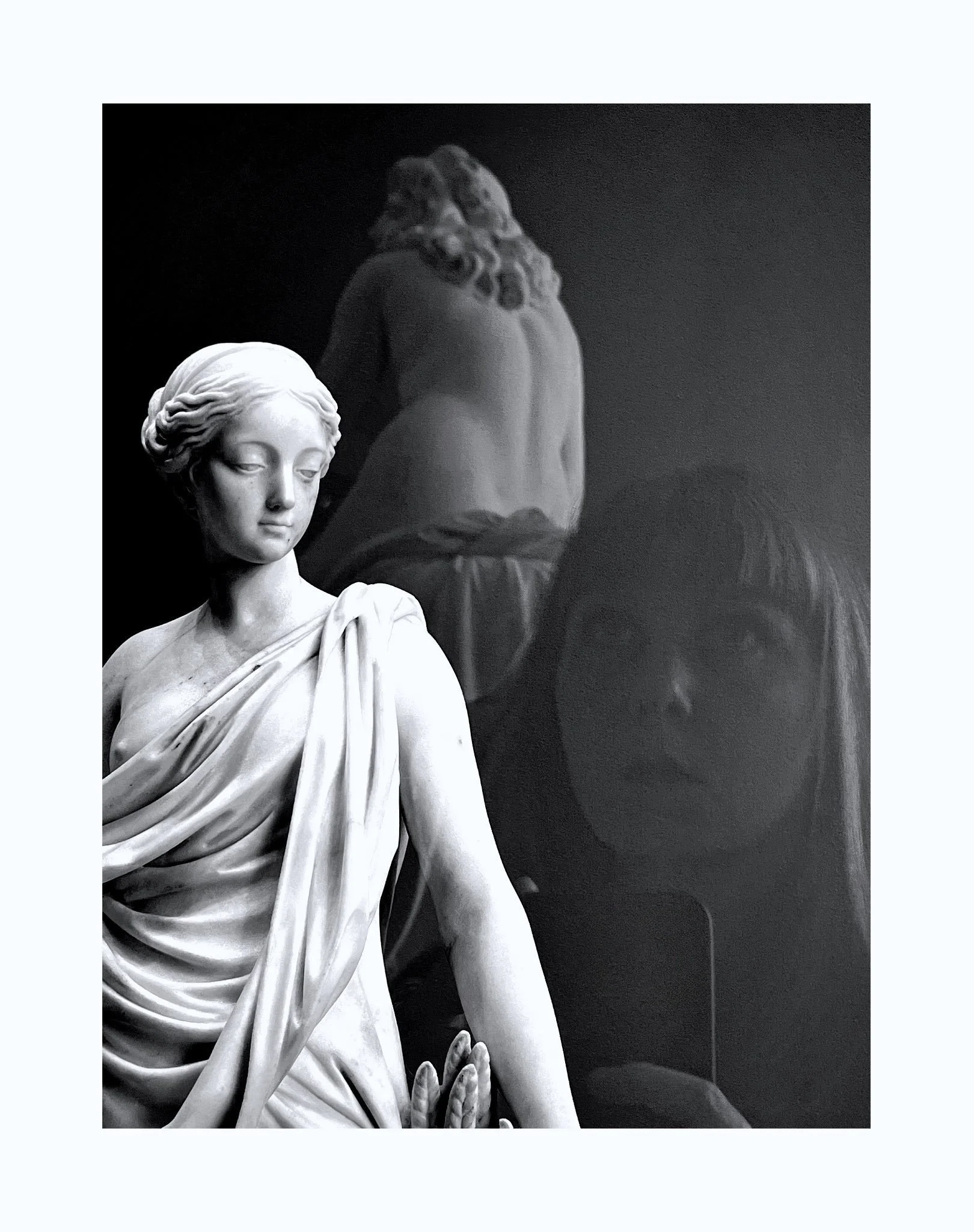 Une sculpture en marbre d'une femme debout, avec des draperies, à côté d'une autre sculpture en arrière-plan et d'un miroir.