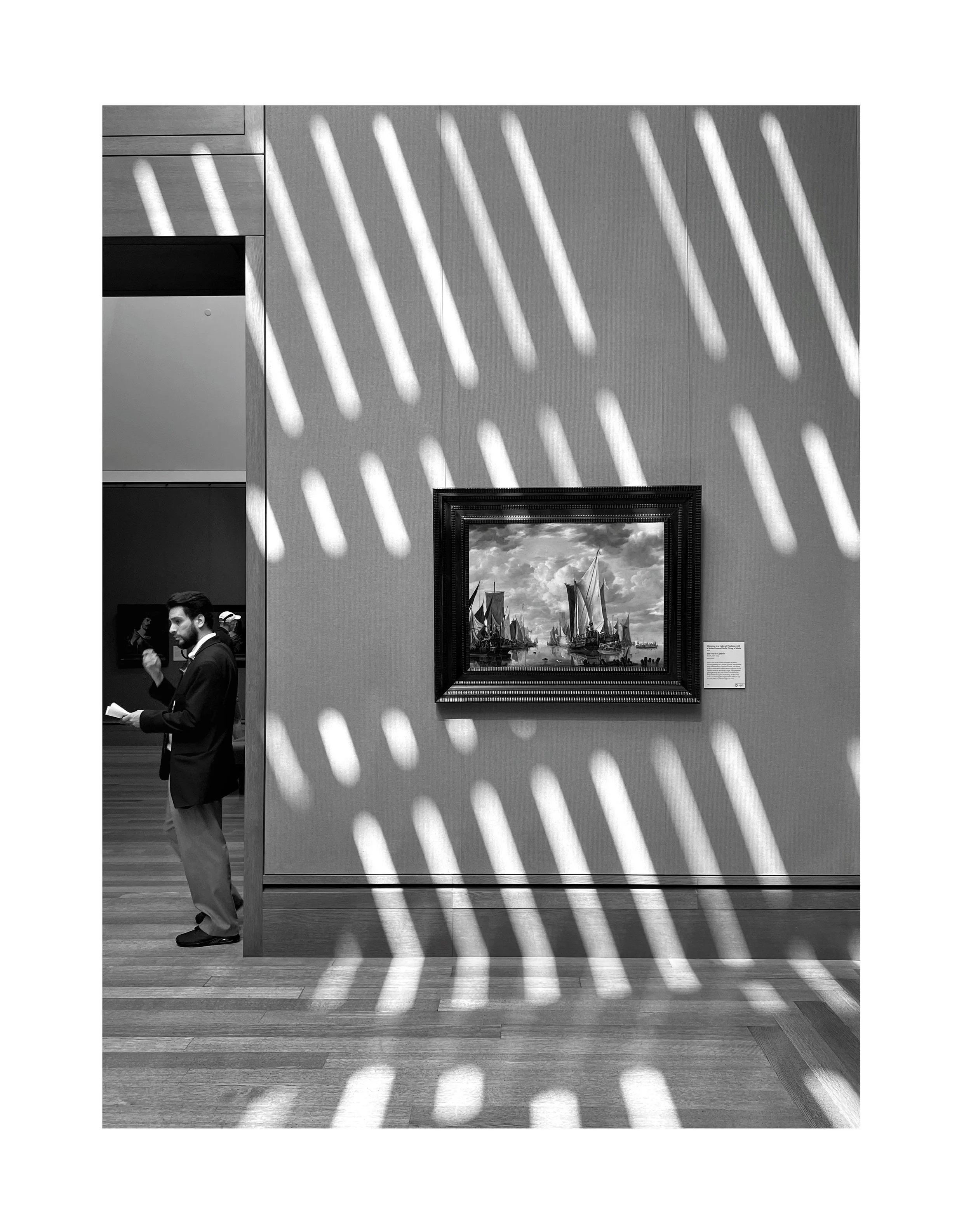 Une œuvre d'art dans un musée, une peinture représentant des voiliers naviguant sur l'eau, entourée d'un cadre décoratif. Des rayures de lumière traversent la pièce en diagonale, créant un motif dans l'espace.