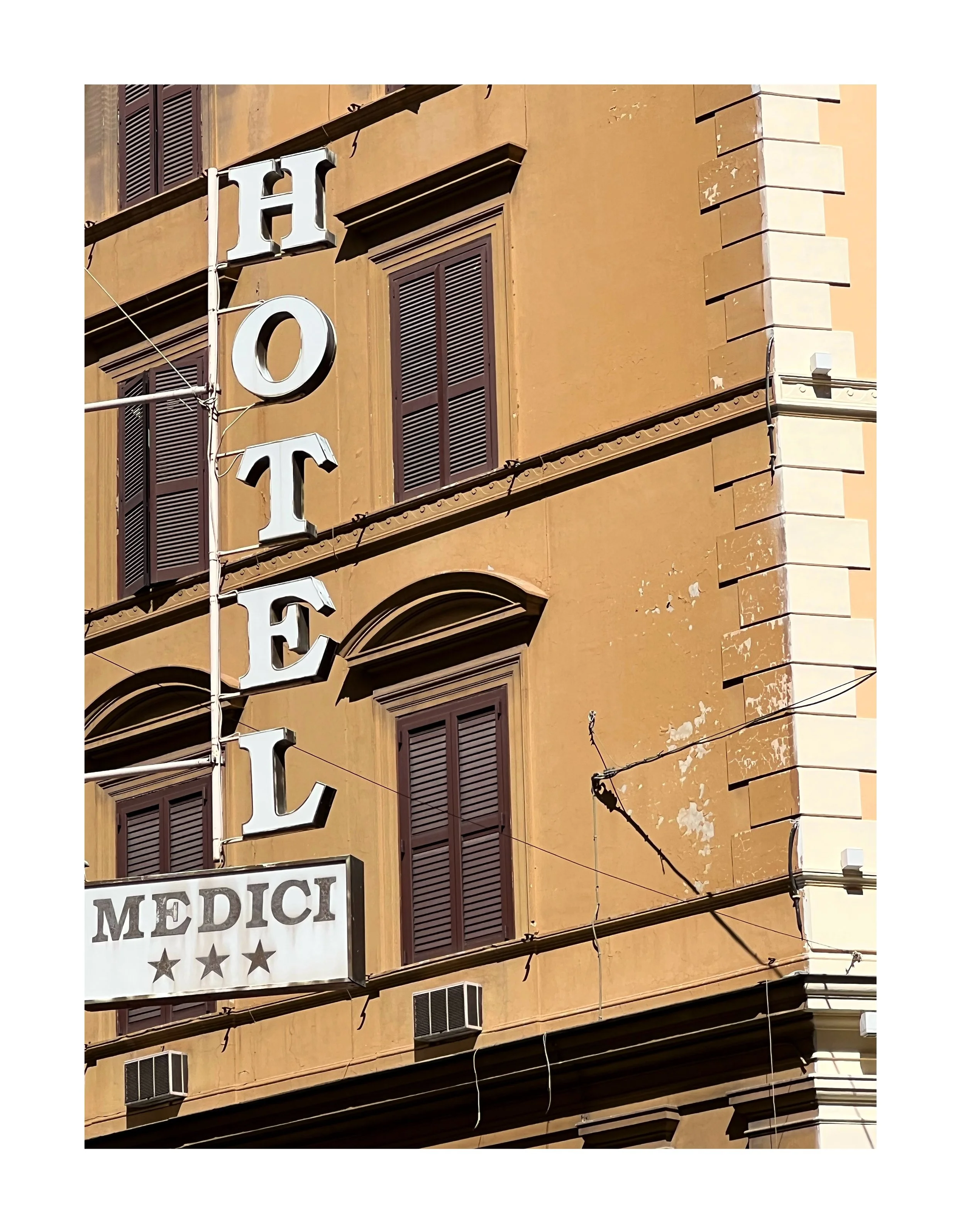 Façade d'un bâtiment avec une enseigne lumineuse indiquant 'HOTEL' et une autre panneau 'MEDICI' avec quatre étoiles, fenêtres à volets en bois.
