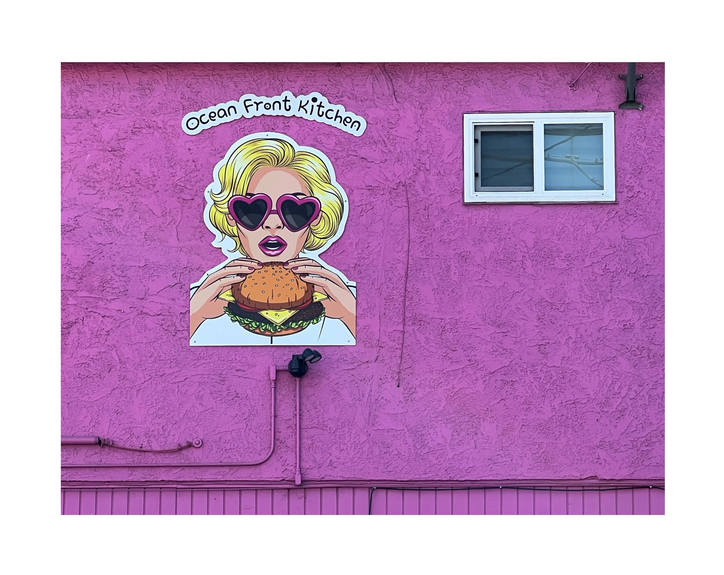 Une peinture murale représentant une femme avec des lunettes en forme de cœur, tenant un hamburger, sur un mur rose avec une fenêtre à droite, dans une enseigne appelée 'Ocean Front Kitchen'.
