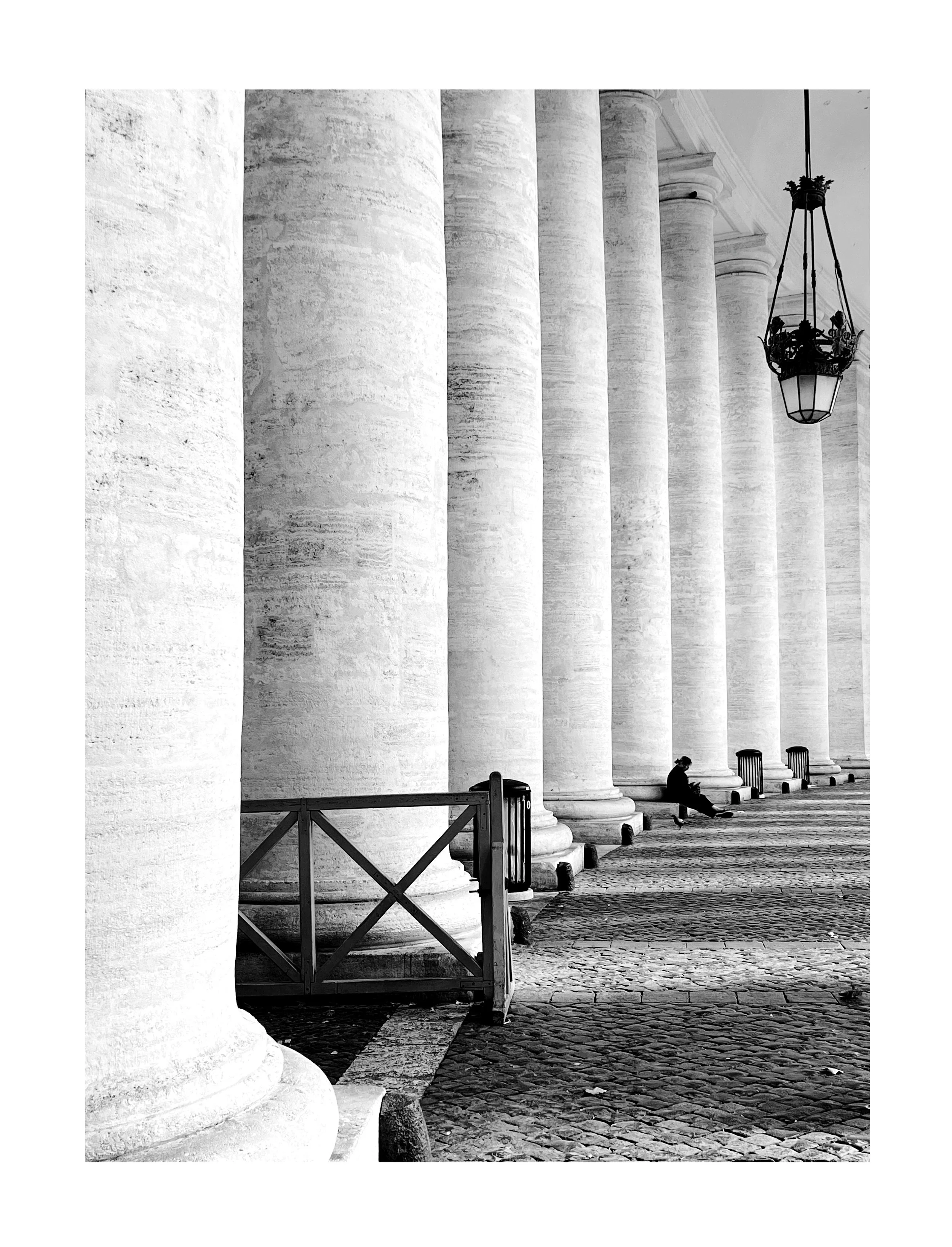 Colonnes en pierre blanche, un lampadaire suspendu, une personne assise à côté des colonnes, sol en pavés.