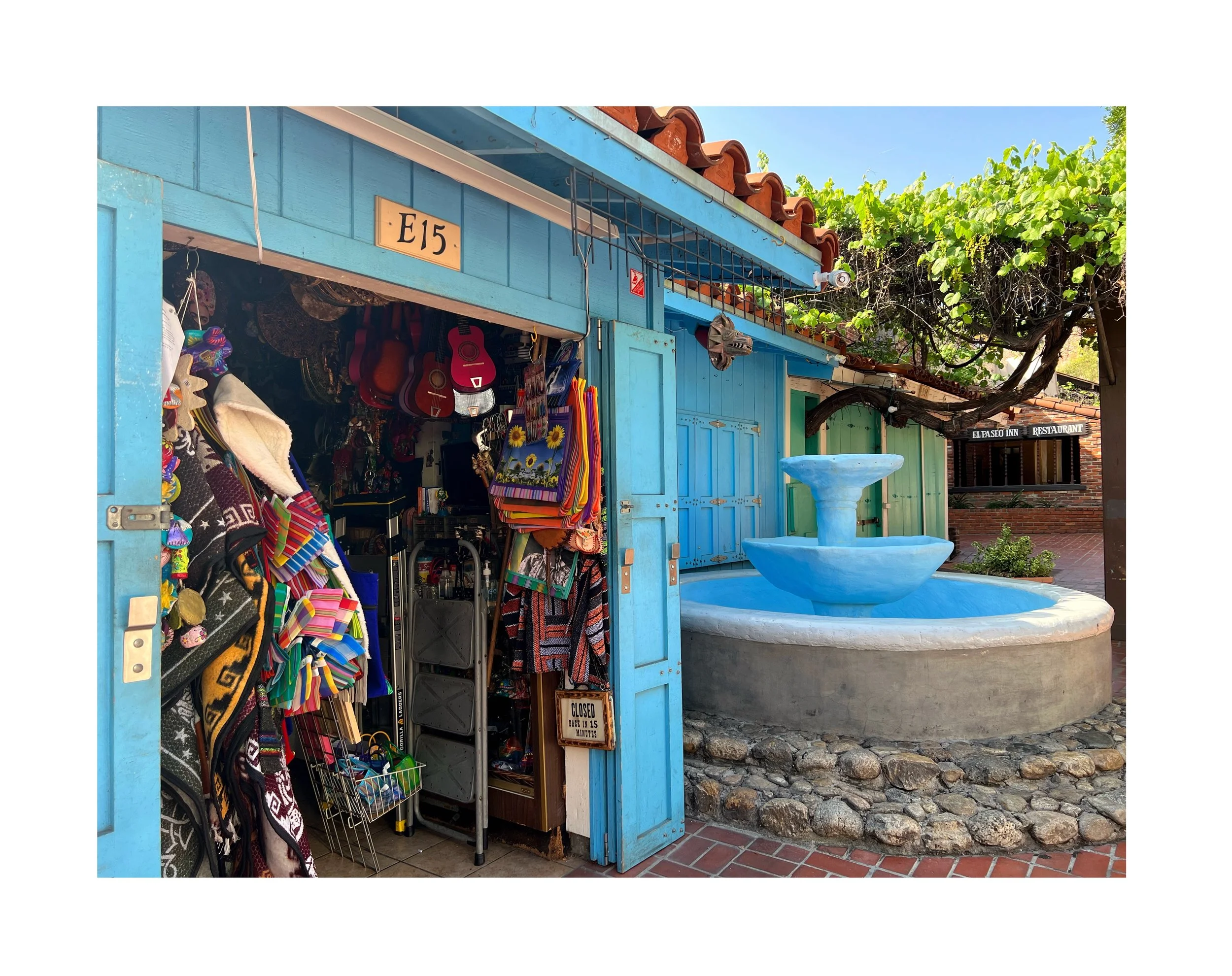 Une petite boutique colorée en bleu avec des souvenirs, des guitares, des sacs et des vêtements suspendus à l'entrée. À côté, une fontaine bleue dans un espace pavé avec des rochers. En arrière-plan, un arbre avec des feuilles vertes et un bâtiment e