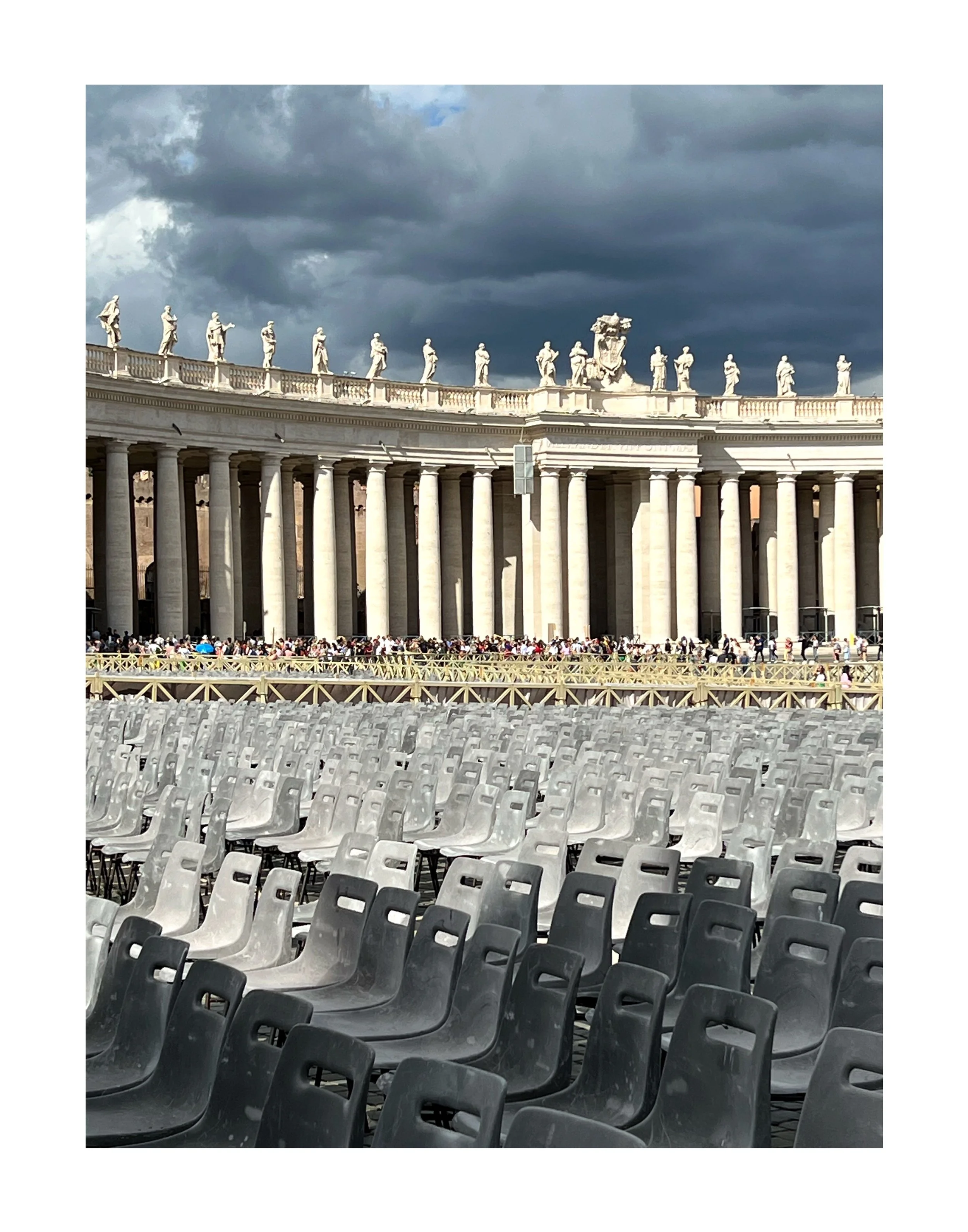Un bâtiment historique avec des colonnes blanches et des statues sur le toit, avec un grand nombre de sièges vides en avant-plan, et un groupe de personnes en arrière-plan, sous un ciel nuageux.