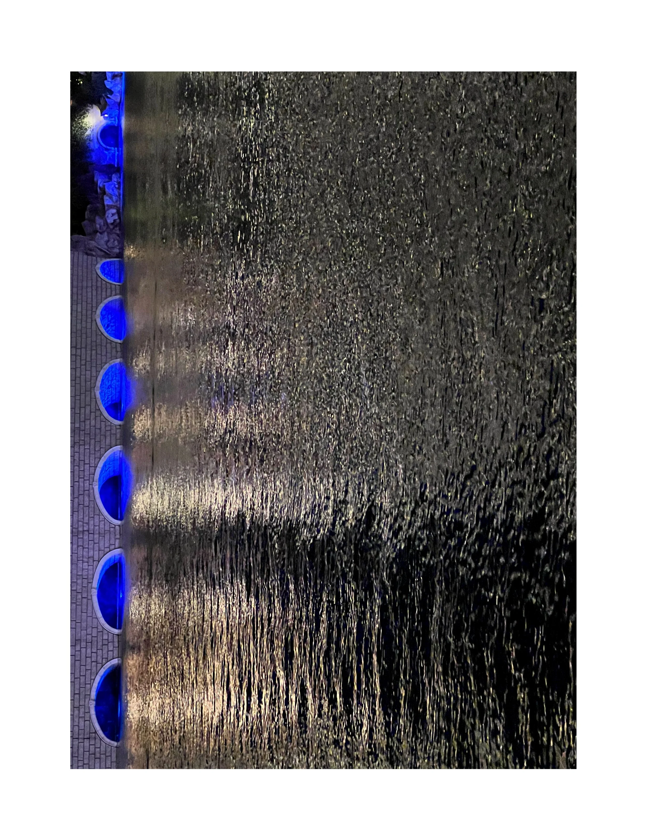 Un mur en briques avec une fontaine en forme de demi-cercle en pierre recouverte d'eau, éclairée par des lumières bleues la nuit.