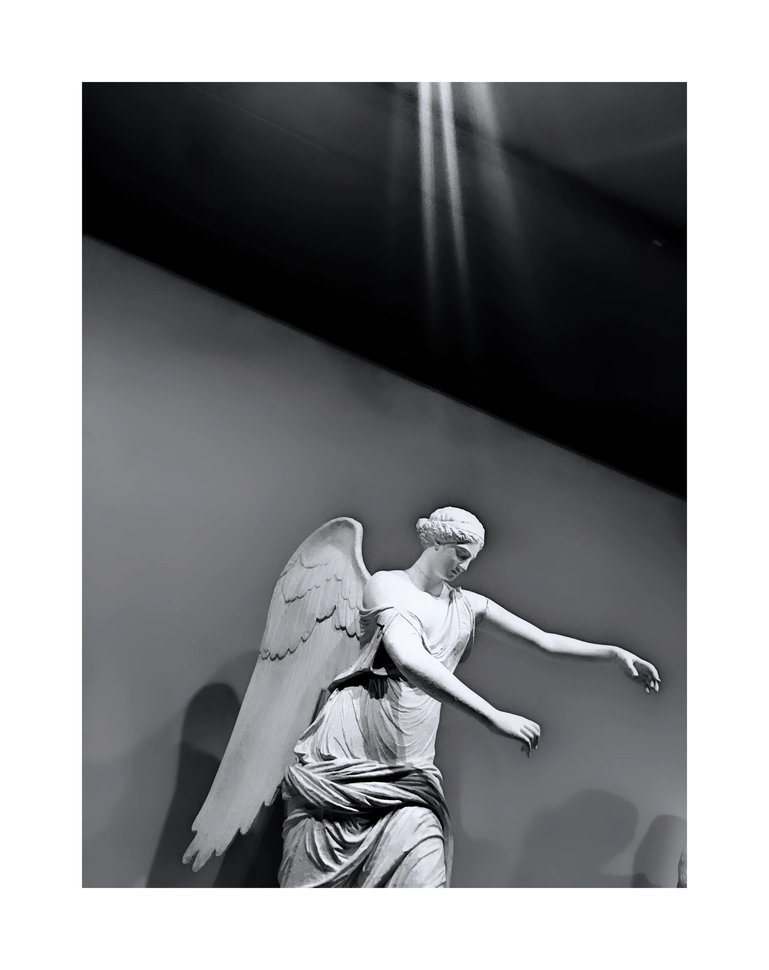 Statue en marbre d'un ange avec des ailes déployées, robe fluide, en mouvement, éclairée par un faisceau lumineux, en noir et blanc.